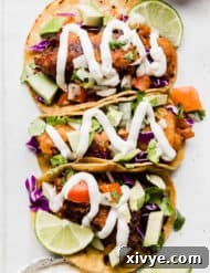 Authentic Baja Fish Tacos topped with white sauce, cabbage, and tomatoes, on a white plate.