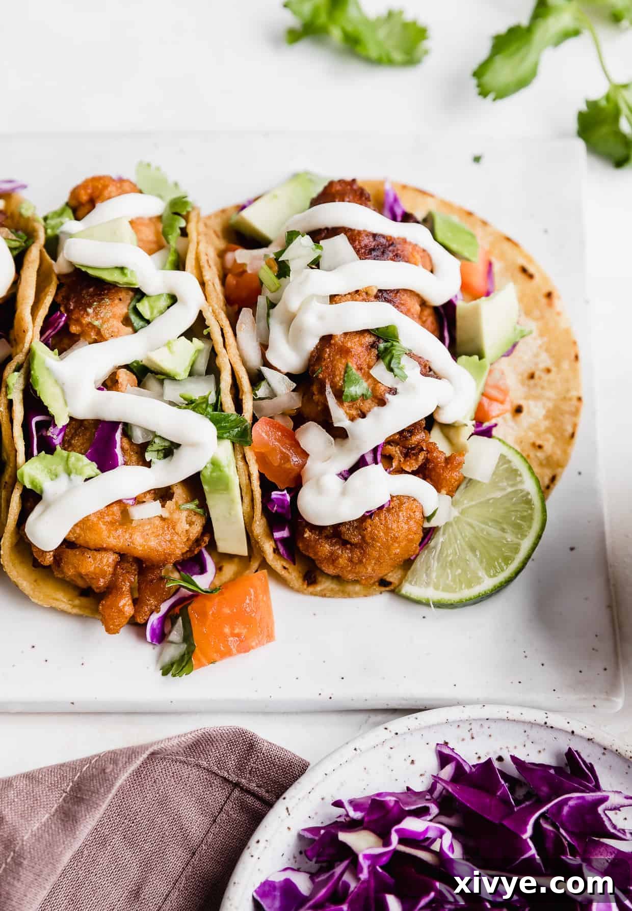 A close up photo of Baja Fish Tacos garnished with purple cabbage, lime wedge, and white sauce. 