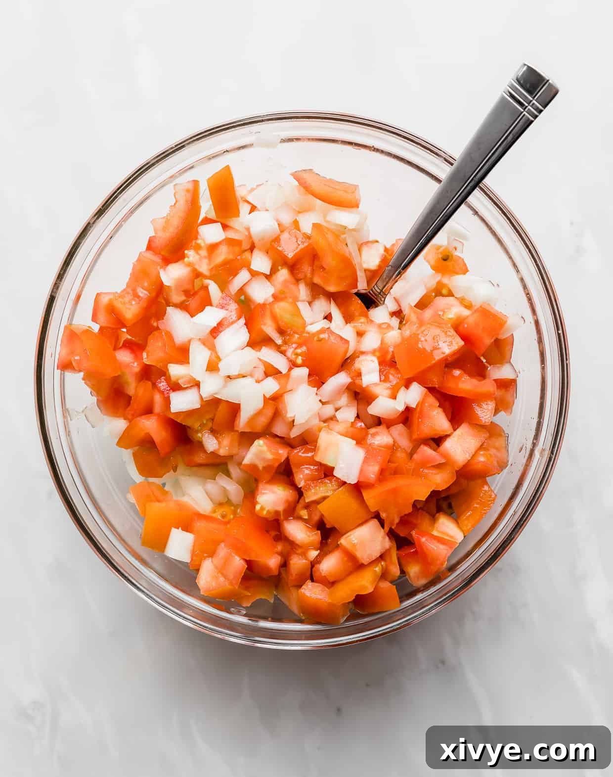 Diced onion and diced tomatoes in a glass bowl against a white background.