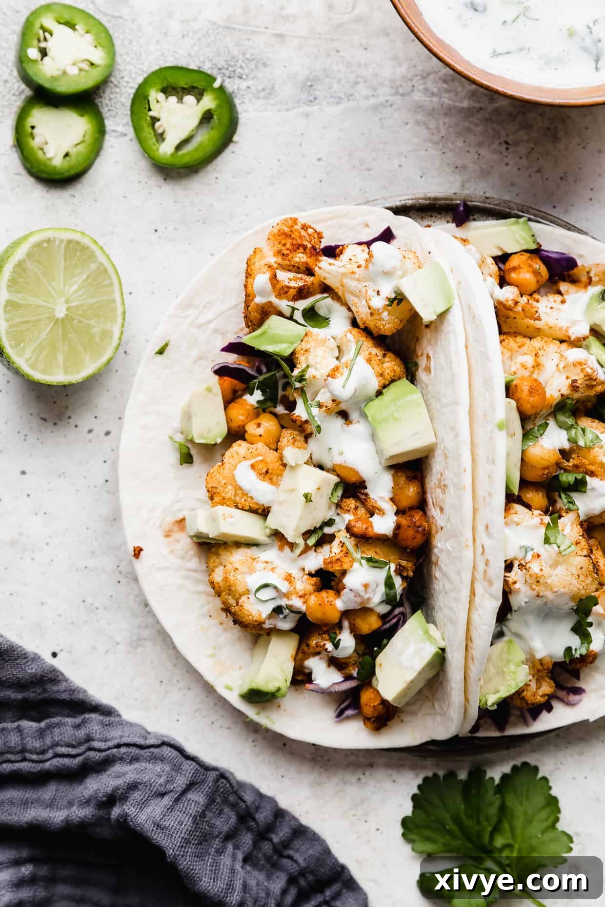 Two flour tortillas filled with a roasted cauliflower and chickpea mixture topped with chopped avocado and a white Crema sauce.