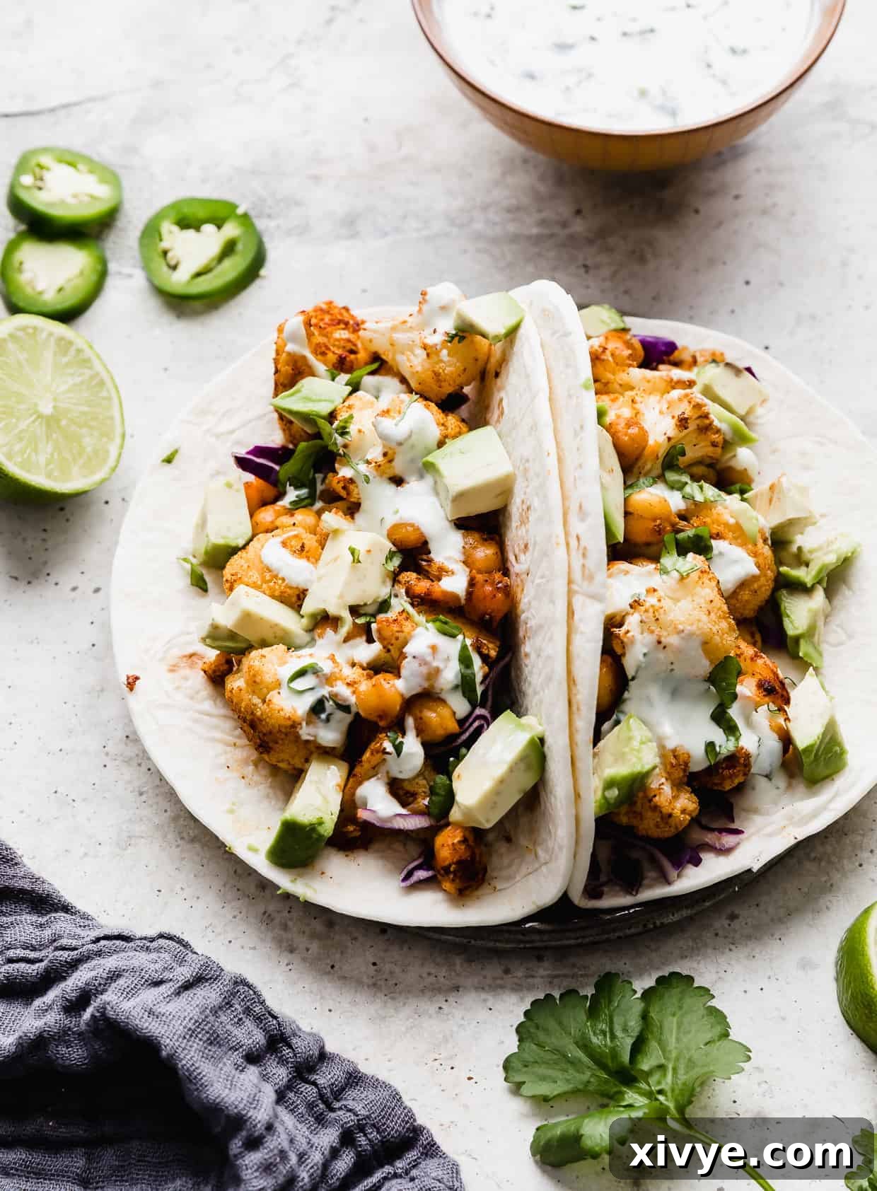 Two cauliflower tacos on a plate. A bowl of white sauce is in the background as well as sliced jalapeños. 