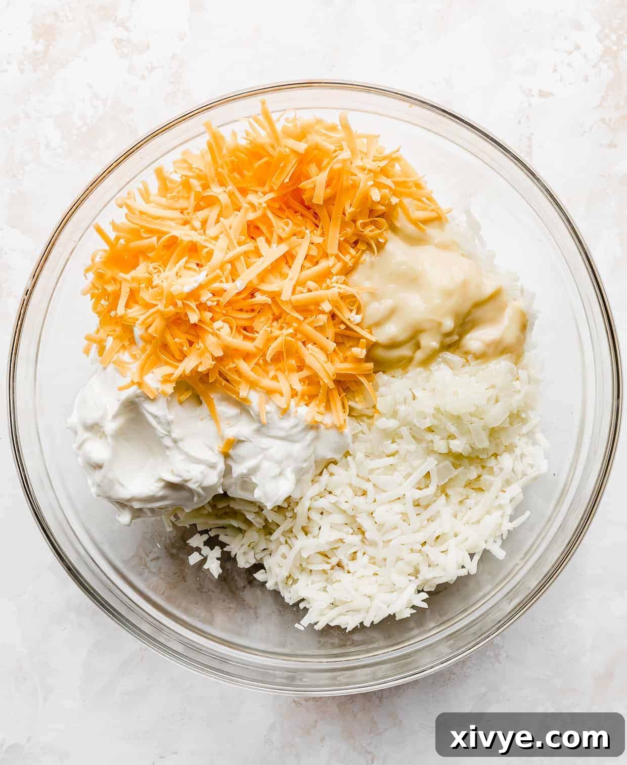 Golden Comfort Bake 4 A large bowl filled with shredded hash browns, cheddar cheese, sour cream, and cream of chicken soup, ready for mixing.