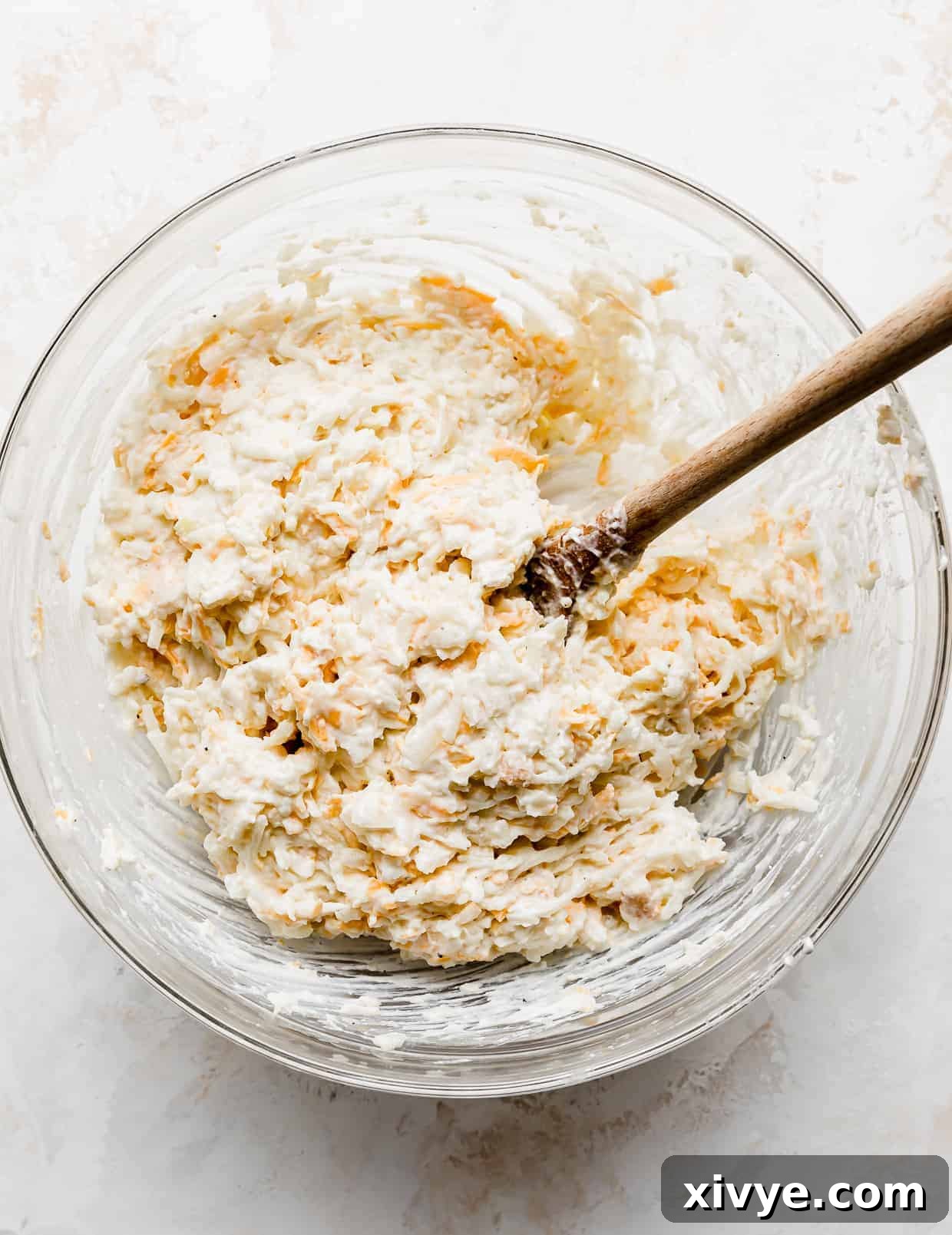 Golden Comfort Bake 5 A large glass bowl with the Funeral Potatoes ingredients all mixed together with a wooden spoon, forming a cohesive casserole base.