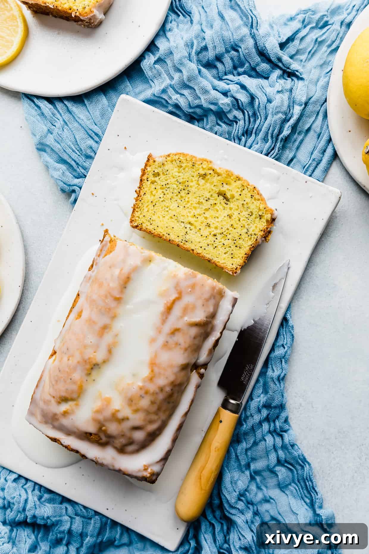 Overhead photo of a freshly baked Lemon Poppy Seed Bread loaf on a white rectangle plate, resting on a blue cloth. The bread is golden brown and generously topped with a white lemon glaze, making it look incredibly inviting and ready to be sliced.