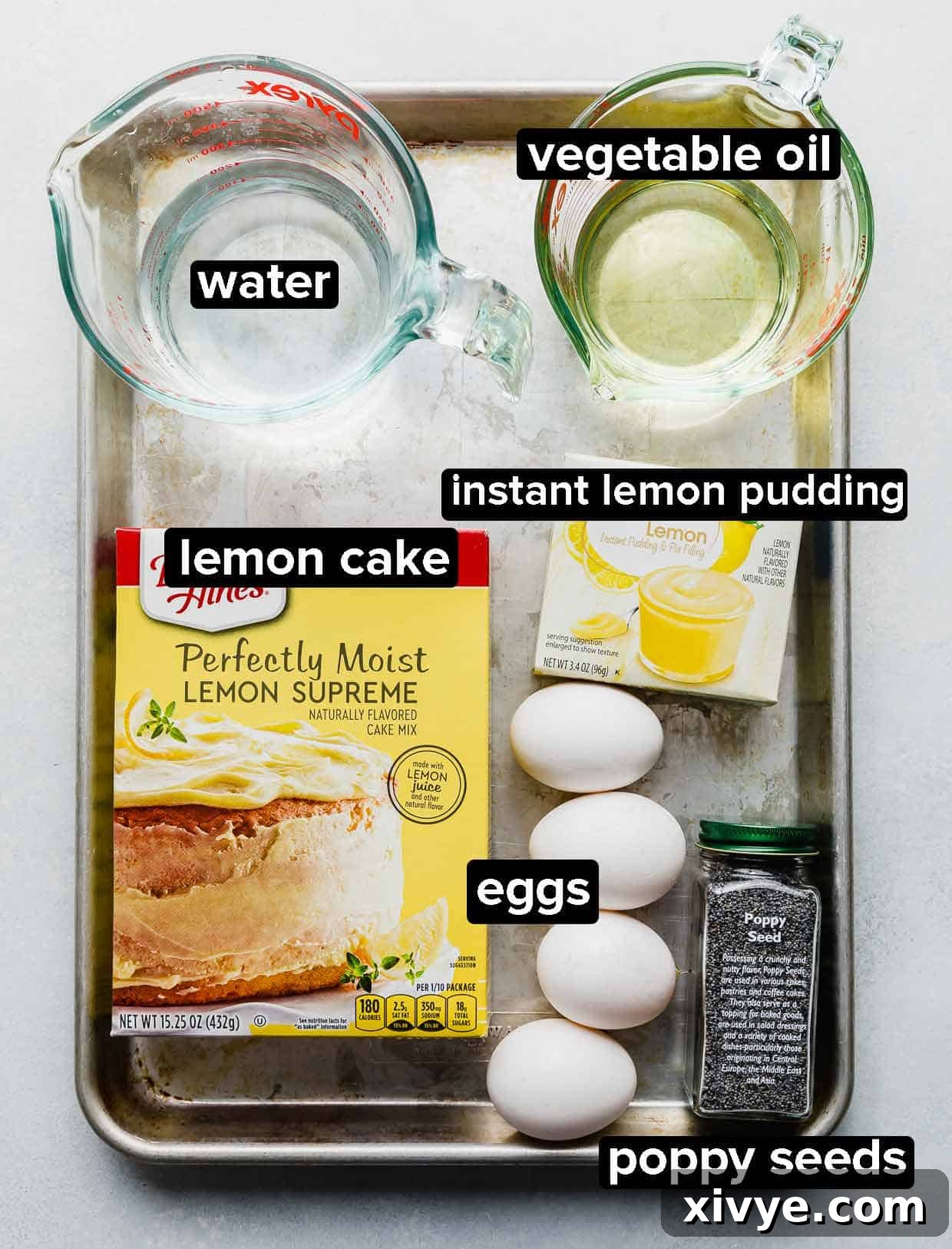 Lemon Poppy Seed Bread ingredients neatly portioned into various glass bowls on a metal baking sheet. Fresh lemons, a box of cake mix, a package of pudding mix, poppy seeds, oil, eggs, and water are visible.
