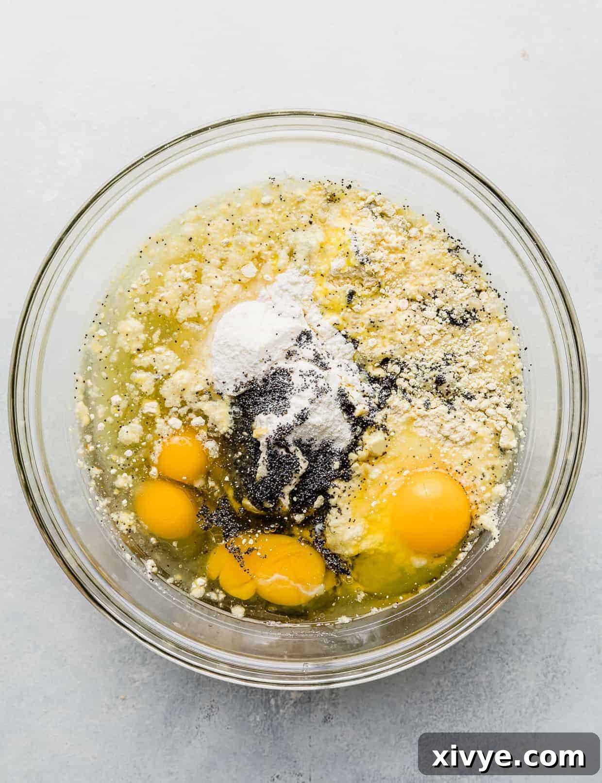 A large glass bowl filled with the unmixed dry lemon cake mix, water, vegetable oil, four cracked eggs, lemon pudding mix powder, and a sprinkle of poppy seeds, waiting to be blended into a smooth batter.