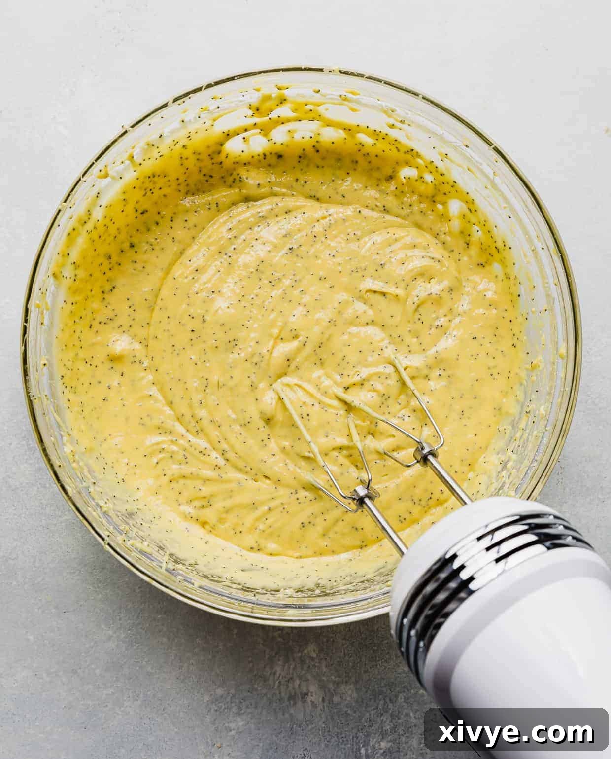 Smooth, pale yellow Lemon Poppy Seed Bread batter, perfectly mixed and uniform, resting in a large glass bowl against a clean white background. A hand mixer is visible, ready for the next step.