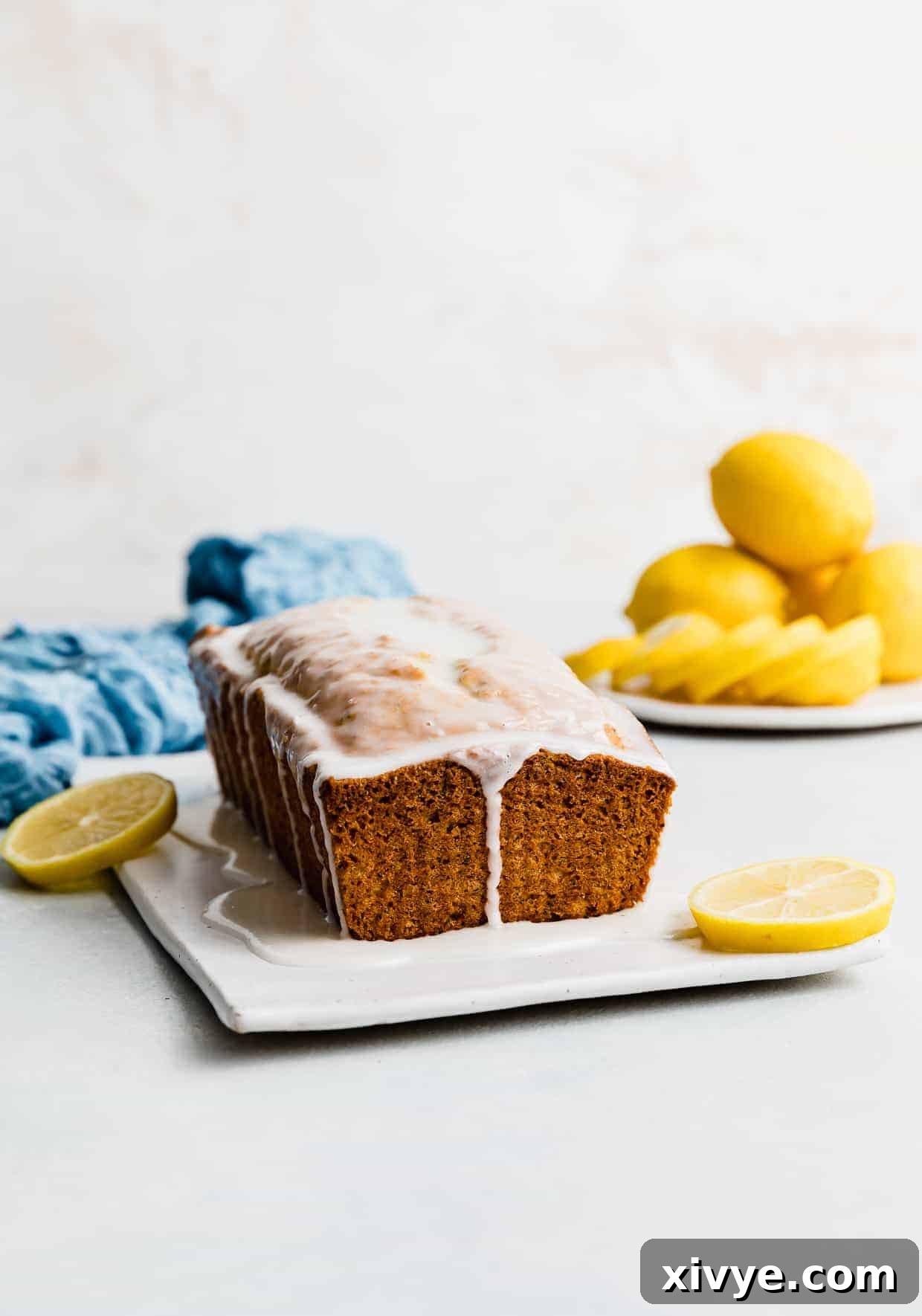 A beautifully glazed loaf of Lemon Poppy Seed Bread resting elegantly on a pristine white background. A small plate stacked with fresh, vibrant yellow lemons sits beside the loaf, enhancing the zesty visual appeal.