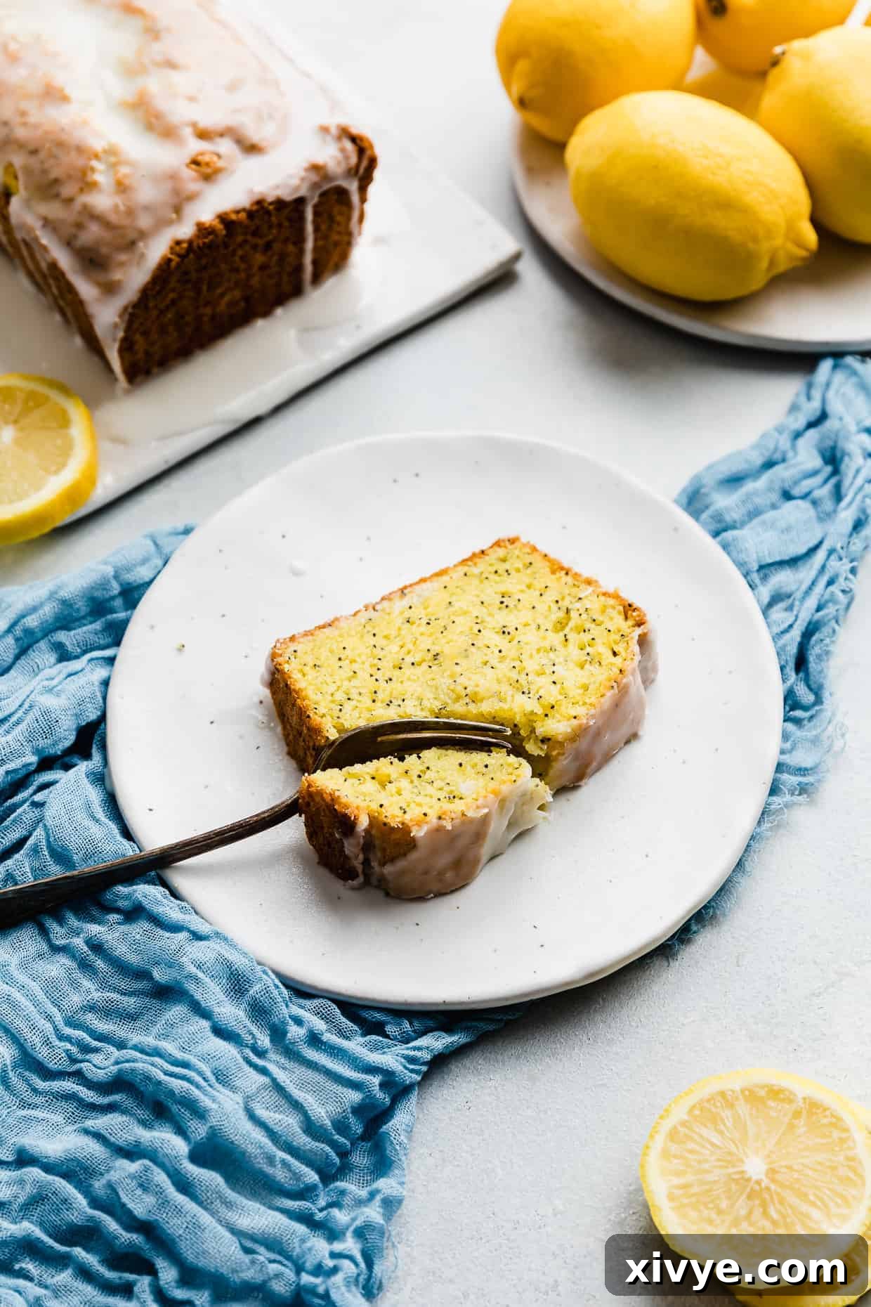 A fork gently cutting into a thick, moist slice of lemon poppy seed bread, glistening with glaze, presented on a simple white plate. The bread's tender crumb and poppy seed specks are clearly visible.