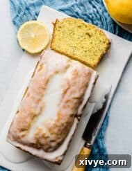 A freshly baked loaf of Lemon Poppy Seed Bread, generously drizzled with a bright white lemon glaze, presented beautifully on a white rectangle plate.
