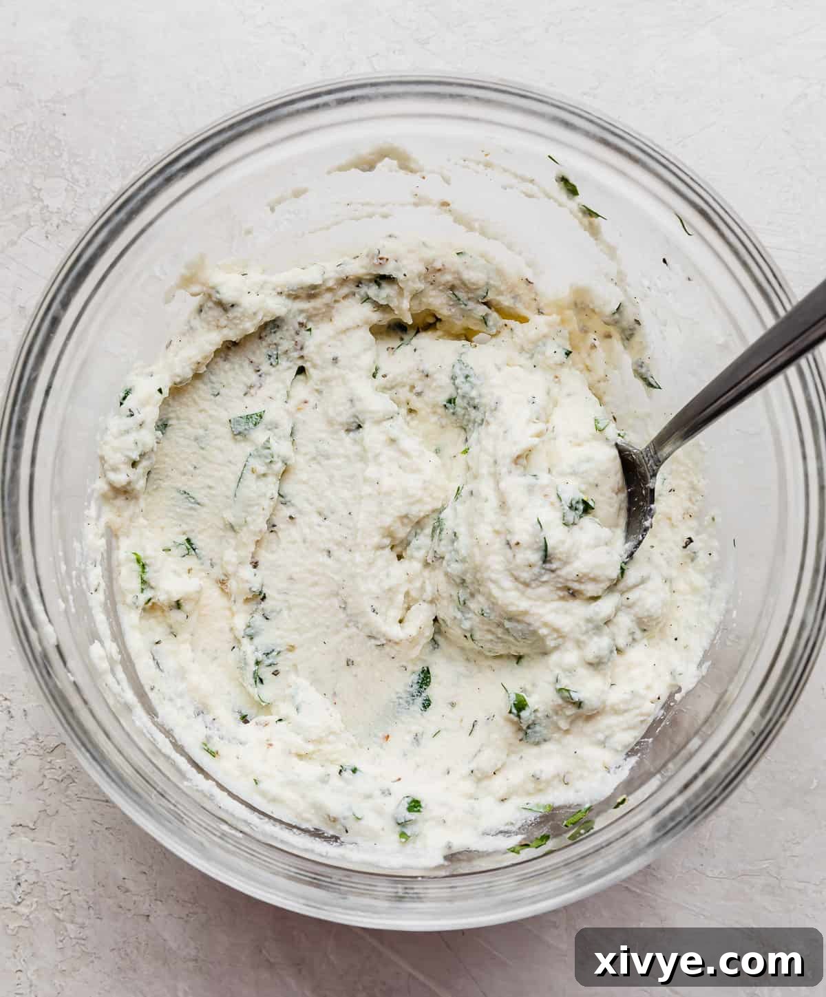 A glass bowl with ricotta mixed with fresh parsley.