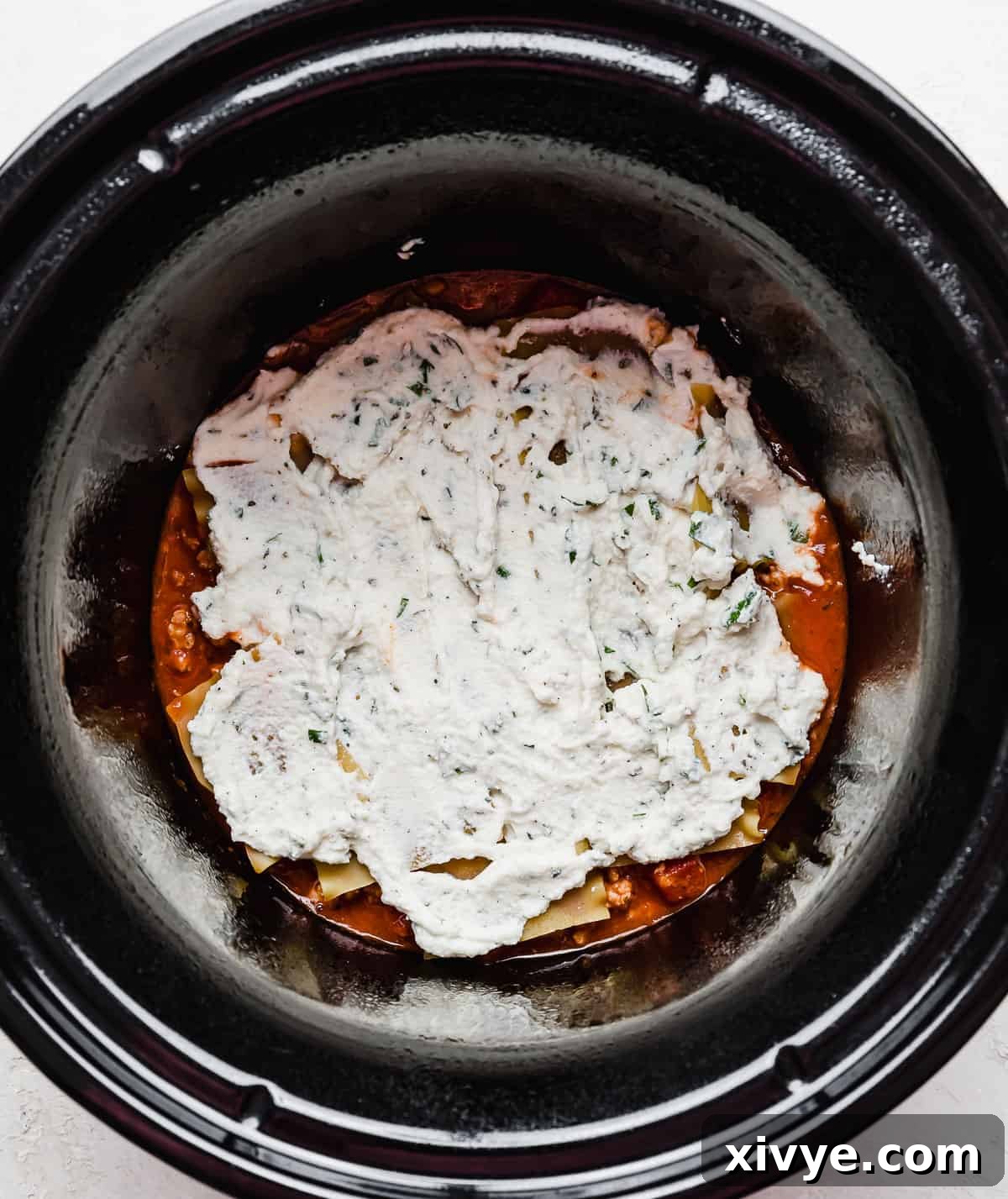 A round slow cooker with ricotta overtop lasagna noodles at the bottom of the crock pot.