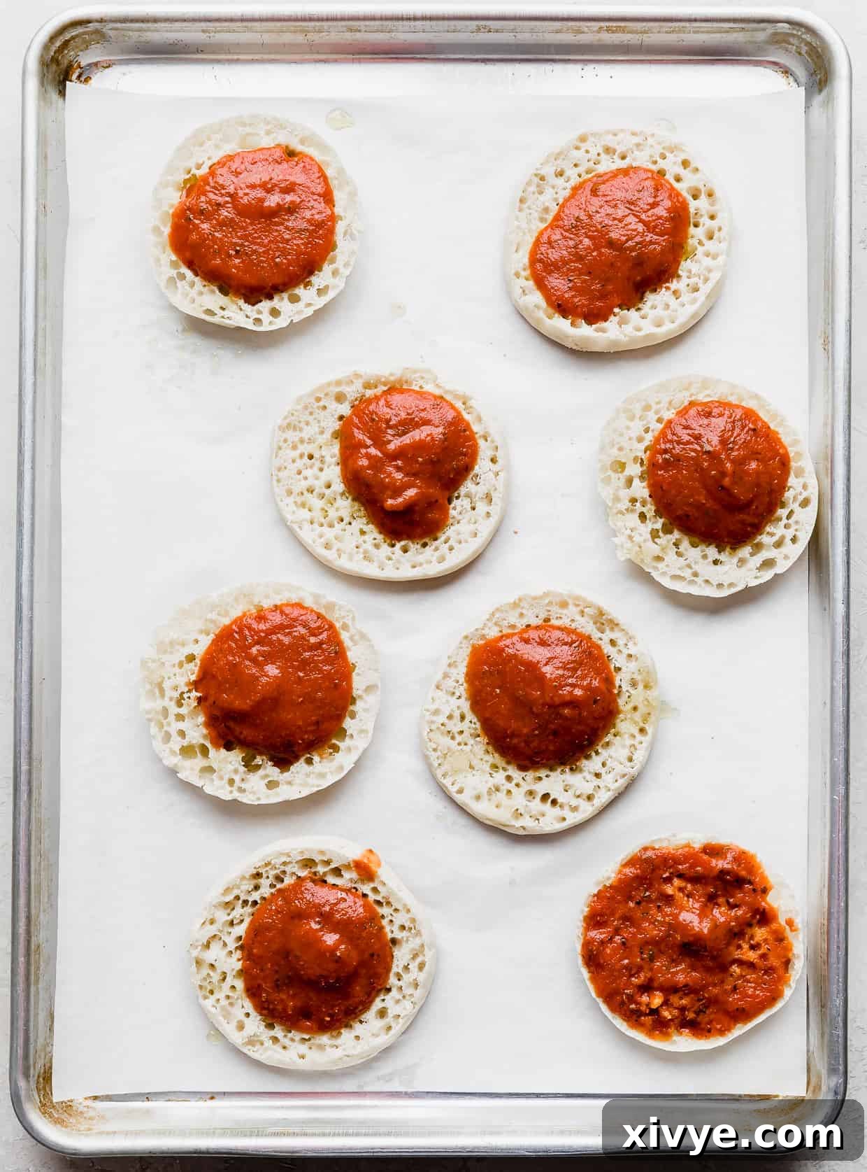 Pizza sauce dolloped on eight English muffin pizza halves, ready to be spread.