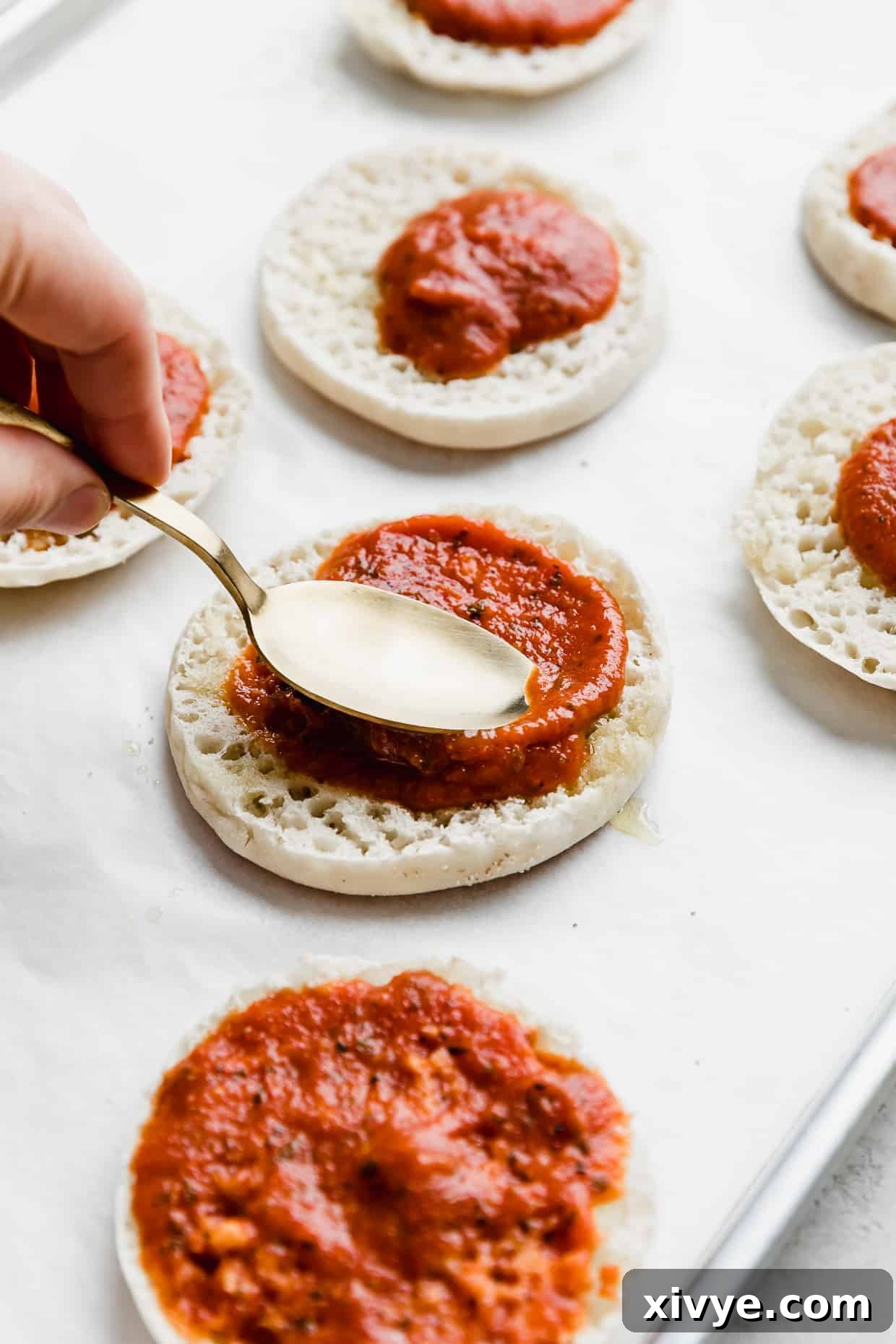 A spoon spreading pizza sauce on a half of an English muffin, ensuring even coverage.
