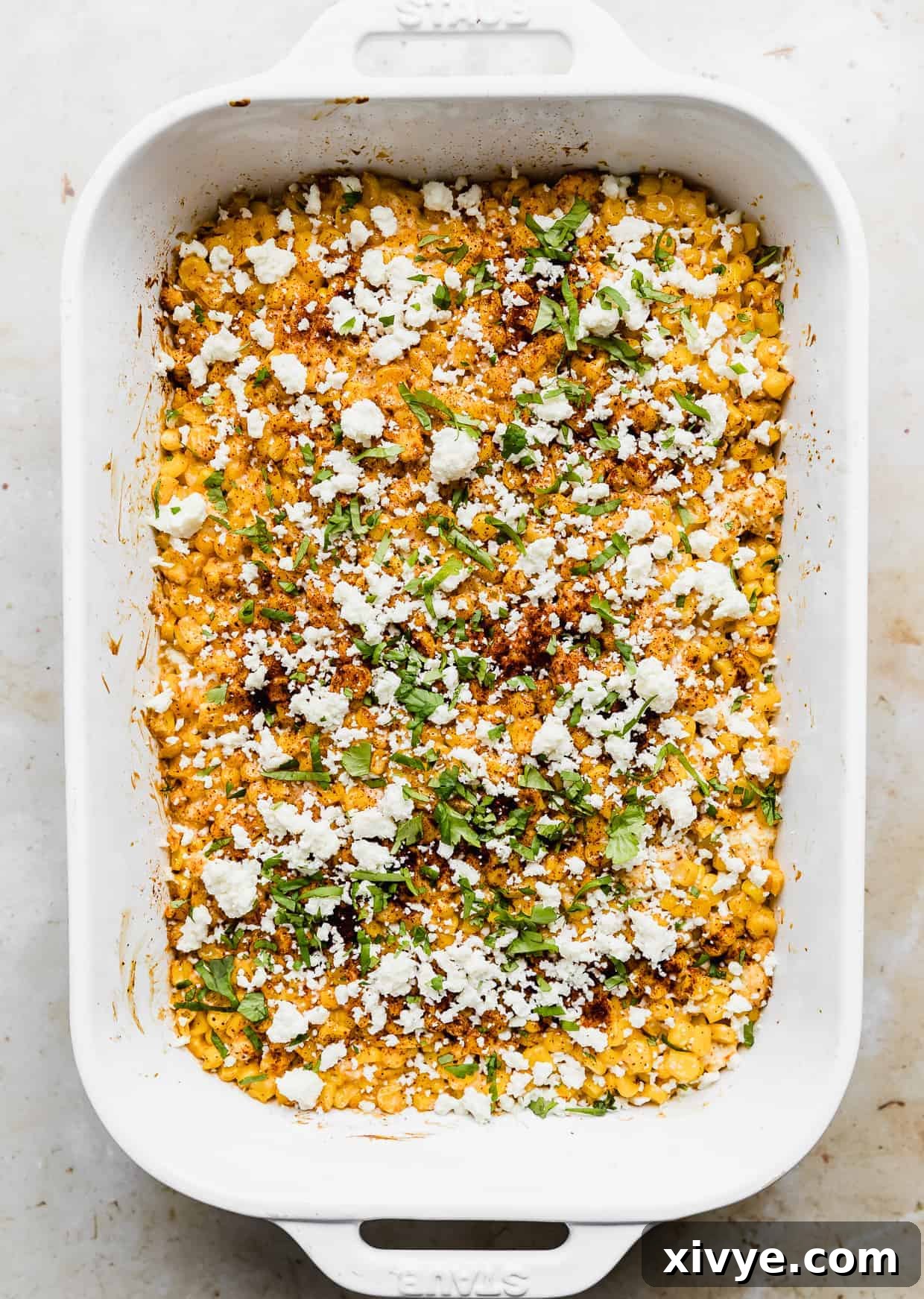 A baked Mexican Street Corn Casserole in a white baking dish, garnished with chopped cilantro, white crumbled cheese, and chili powder.