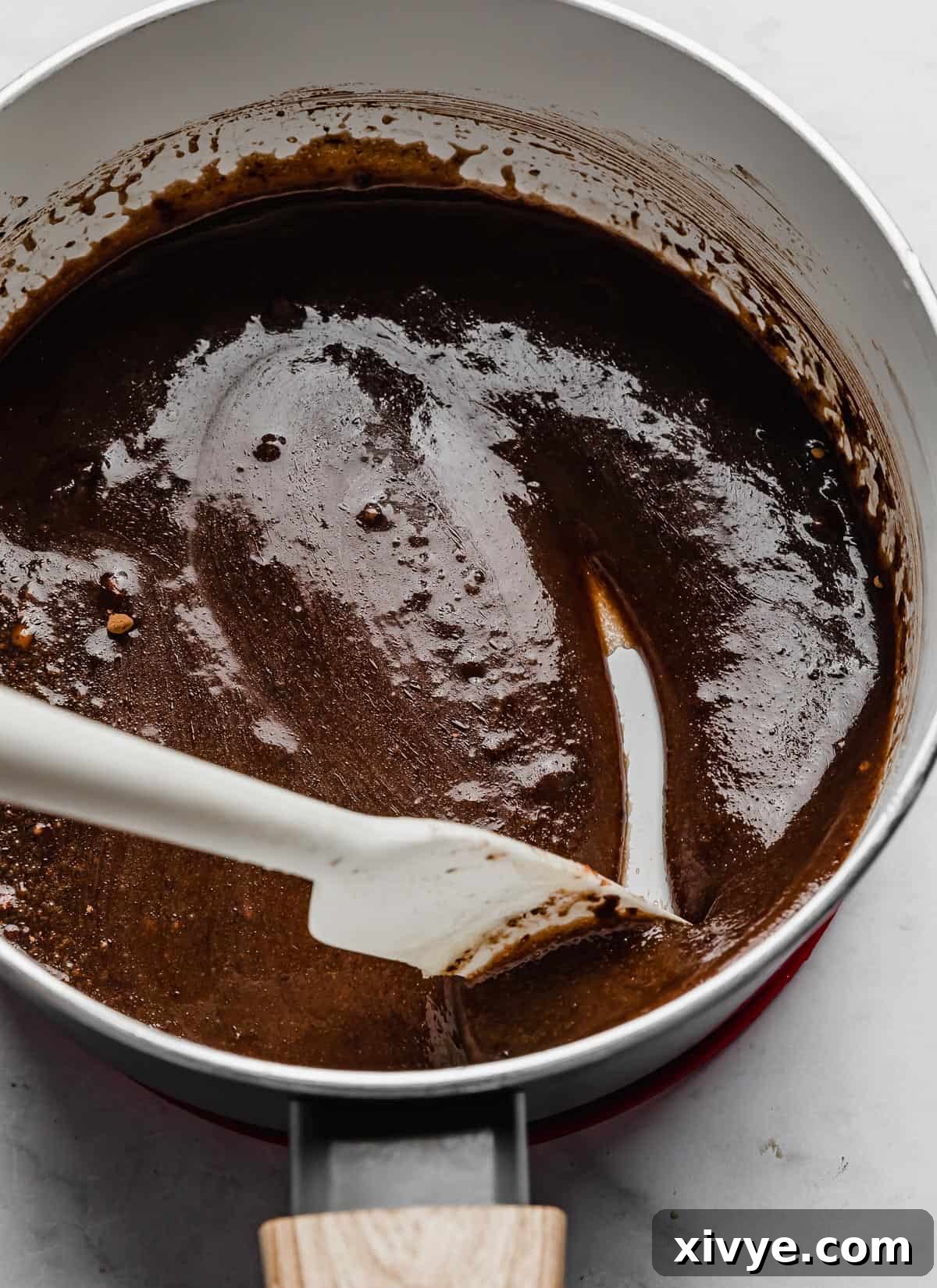 A white spatula stirring a rich chocolate mixture in a white pot, reaching a smooth consistency.