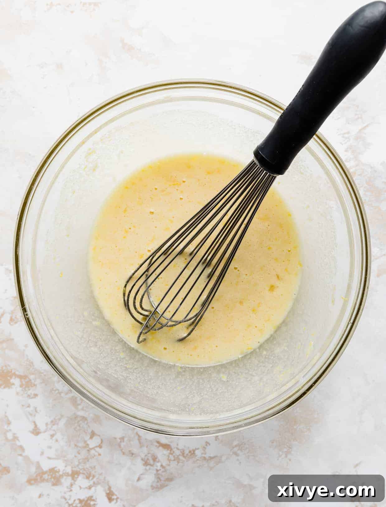 A glass bowl containing a vibrant yellow lemon bars filling, with a whisk resting inside, showing the creamy texture.