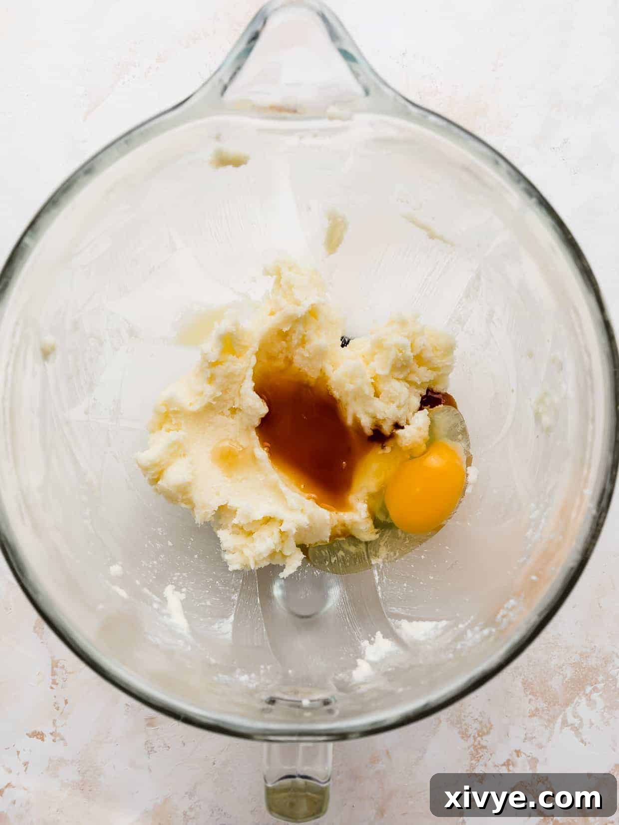 A glass bowl with creamed butter and sugar, vanilla extract, and a cracked egg in it, against a white background.