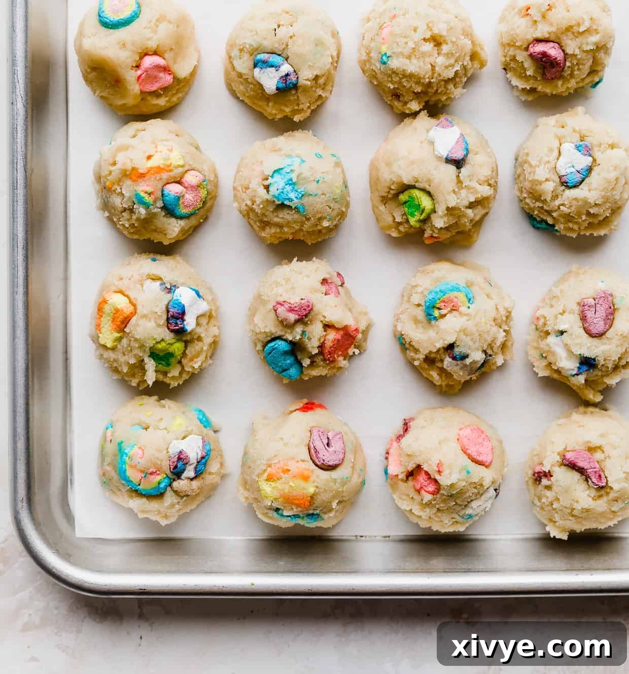 A white parchment lined baking sheet with balls of sugar cookie dough balls topped with lucky charms marshmallows on the tops. 