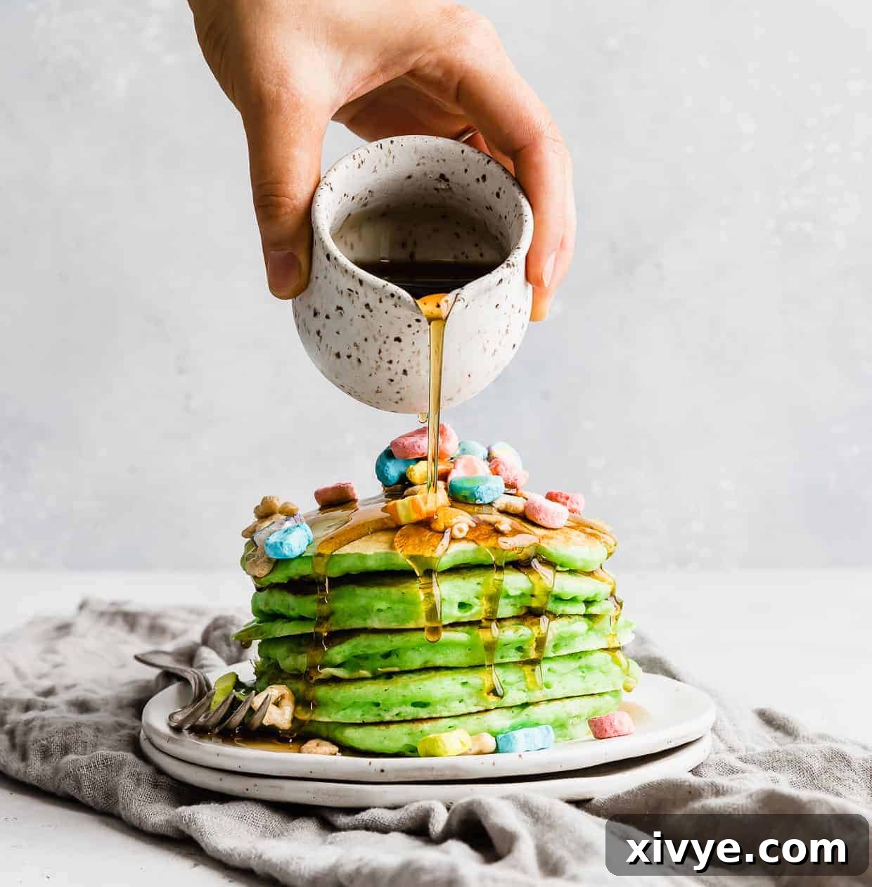 A hand pouring maple syrup over a stack of green pancakes on a white plate. 