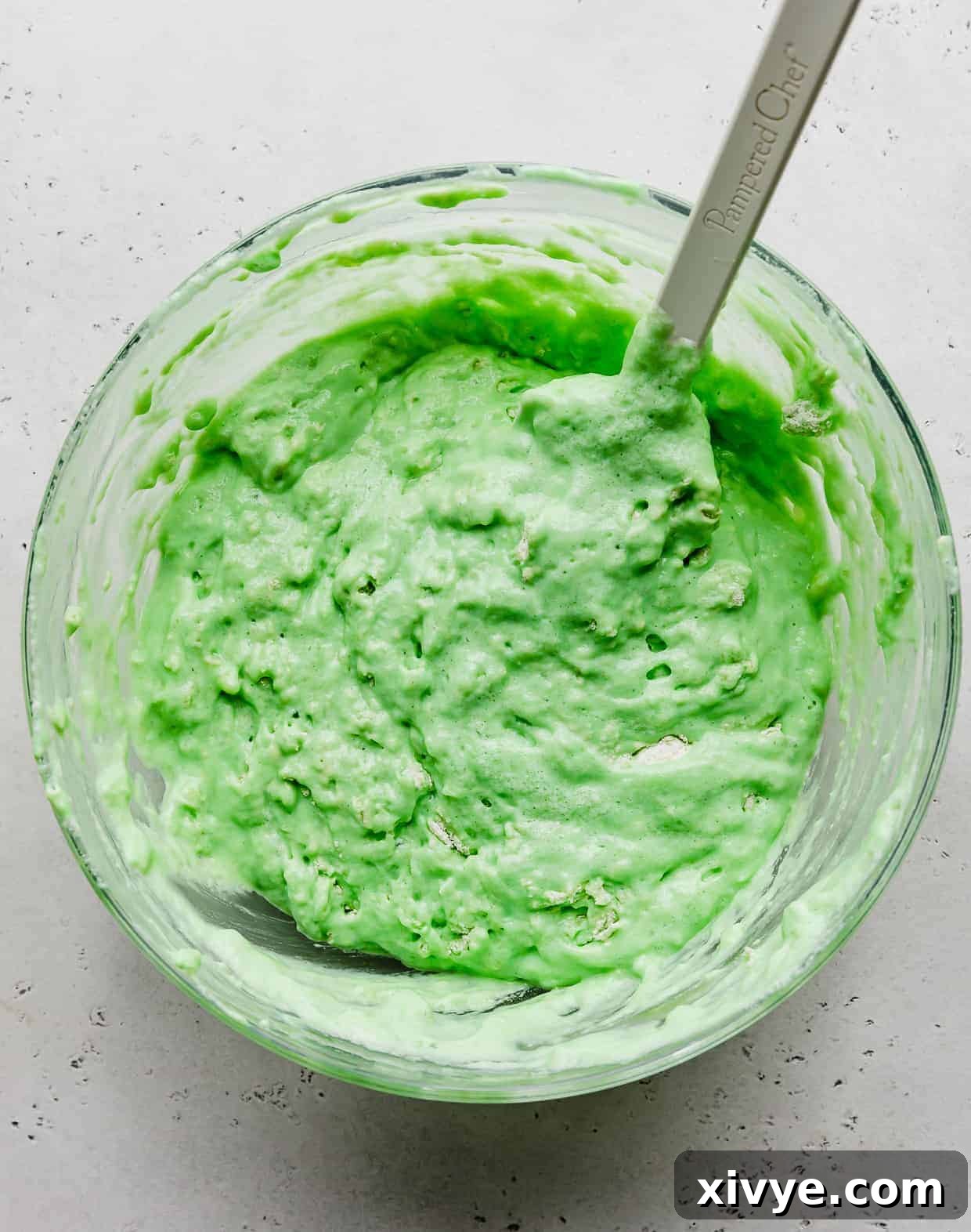 A thick green pancake batter in a glass bowl against a light gray background. 