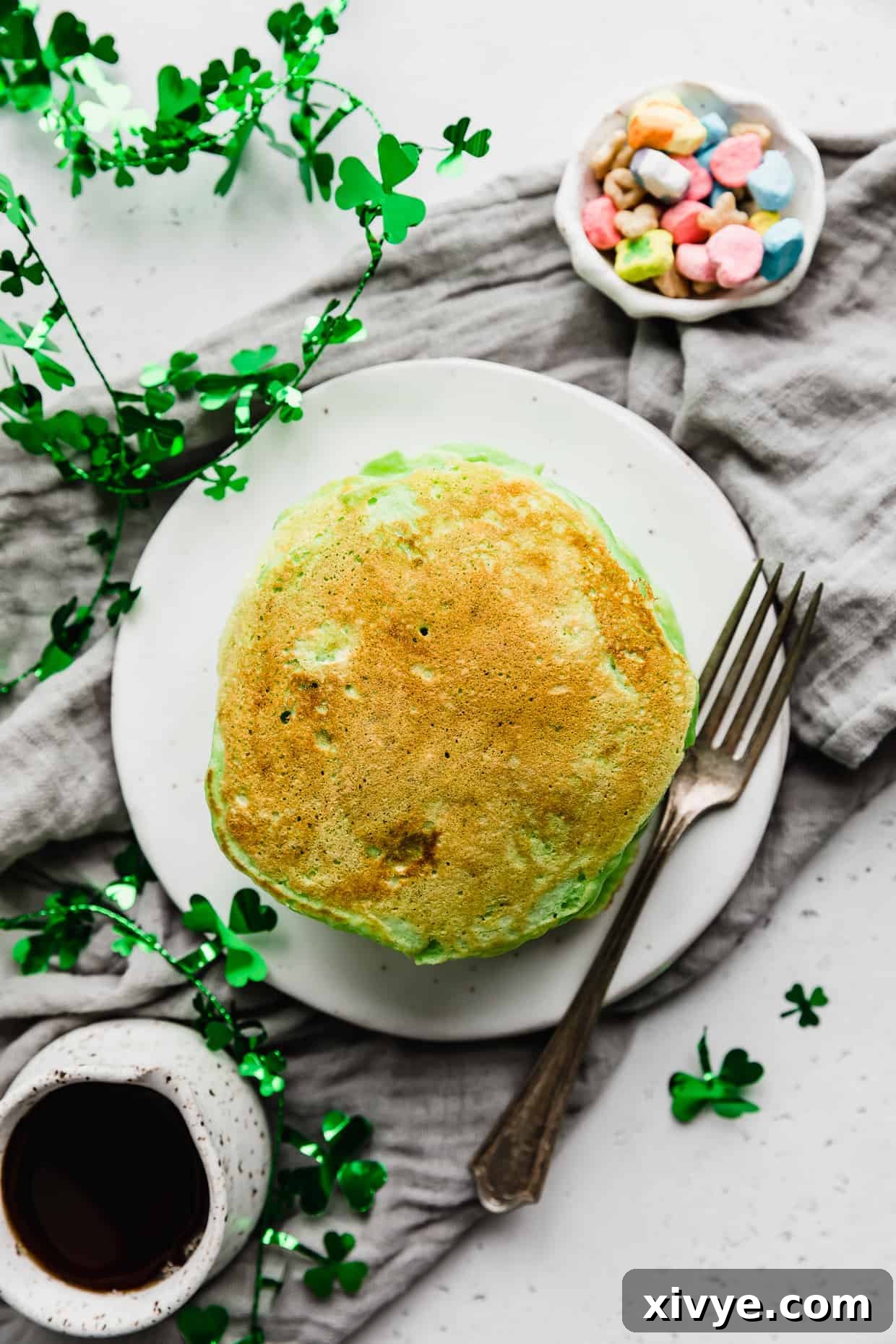 Overhead photo of green pancakes on a white plate with lucky charms marshmallows in a small bowl next to the pancakes. 