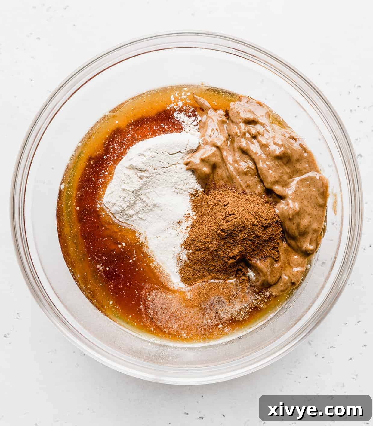 A glass bowl full of ingredients used to make Snickerdoodle Energy Bites; almond butter, maple syrup, protein powder, cinnamon, and kodiak cakes mix. 