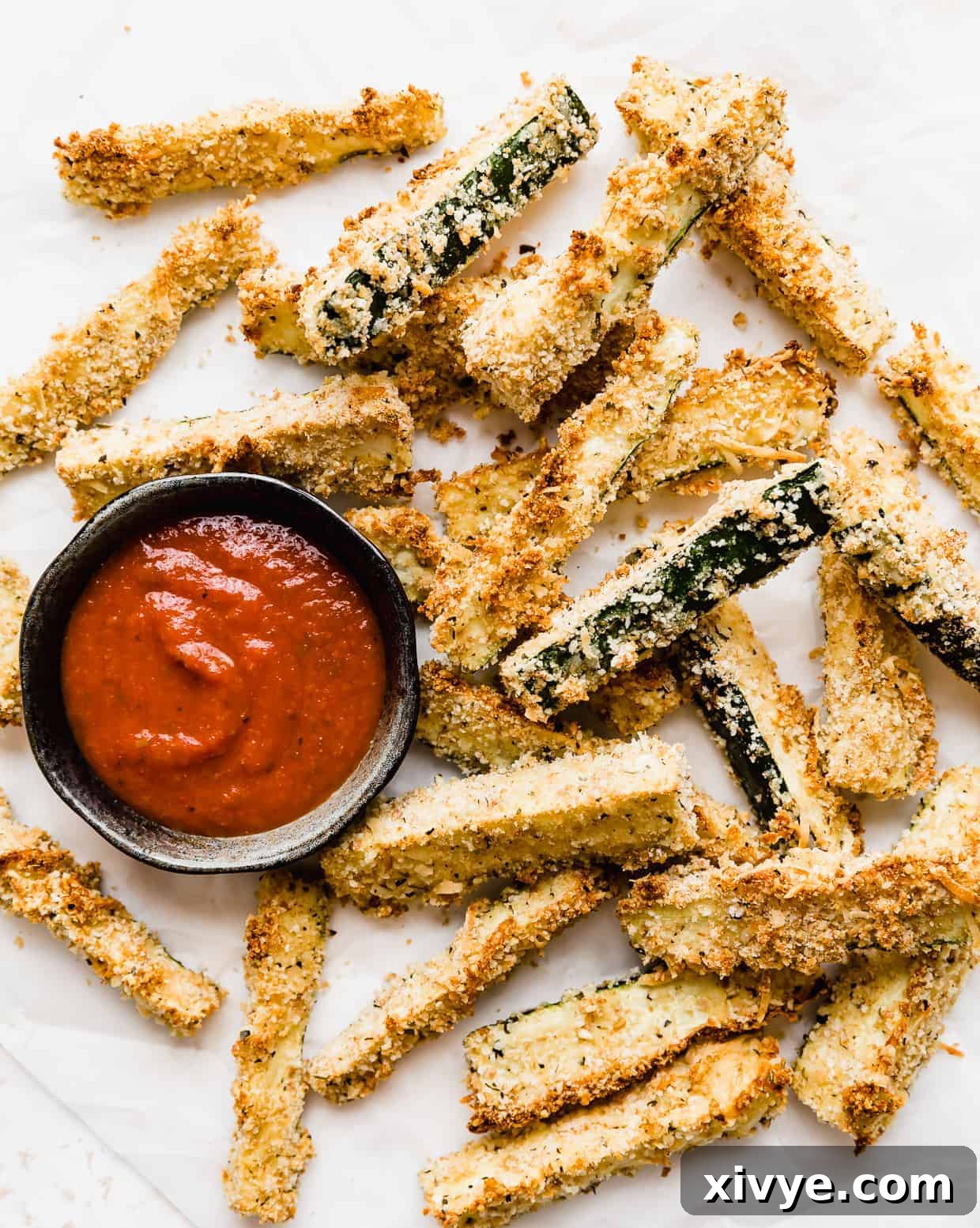 Baked Zucchini Fries on a white parchment paper with a small bowl of marinara sauce next to the fries. 