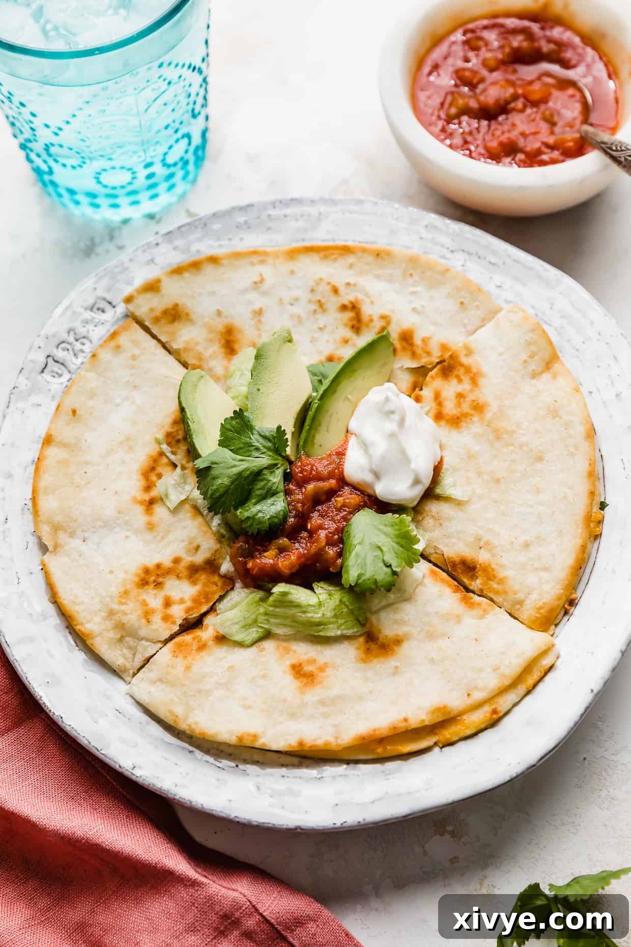 A cheese quesadilla on a white plate topped with avocado, salsa, sour cream, and shredded lettuce.