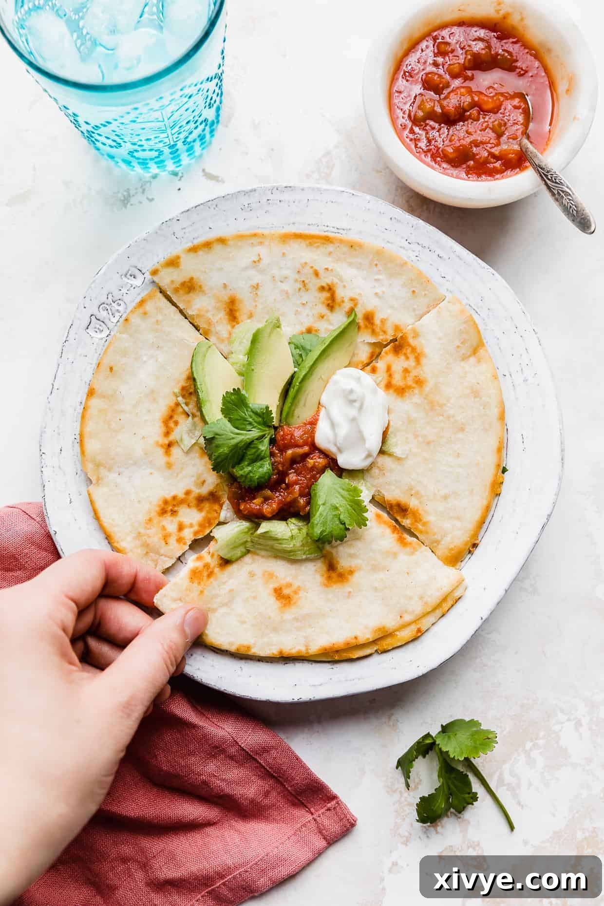 A hand grabbing a slice of a golden brown cheese quesadilla on a white plate that has been topped with avocado, sour cream, and salsa.
