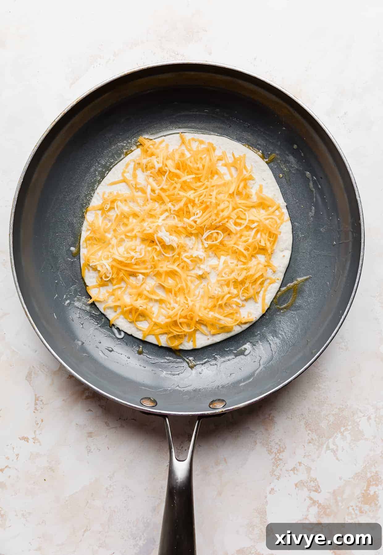 A frying pan with a quesadilla in it that has been topped with shredded cheddar cheese.