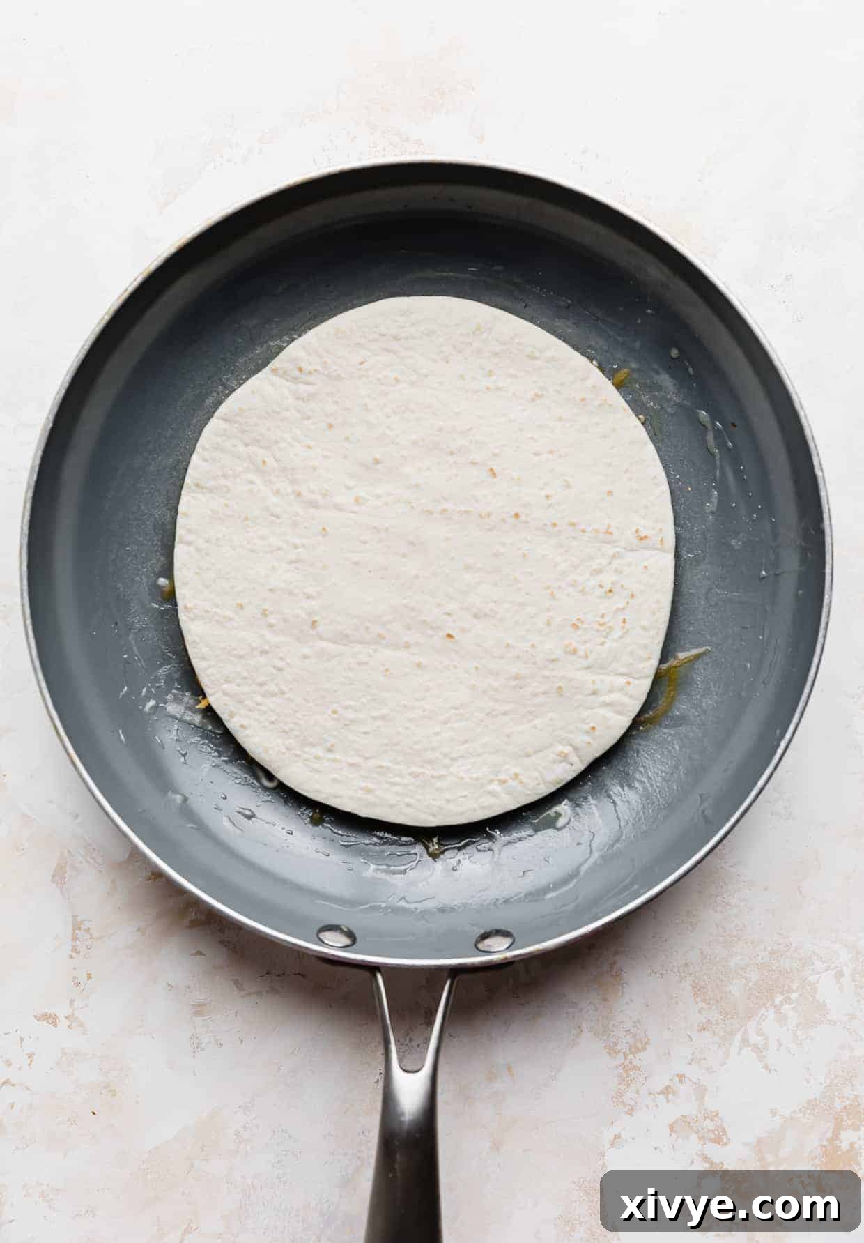 A tortilla in a gray frying pan against a white and cream colored background.