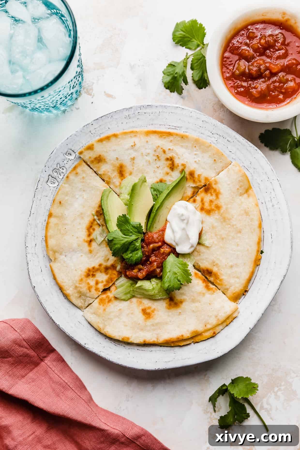 A cheese quesadilla on a white plate that has been cut into 4 wedges, topped with salsa, avocado, sour cream, and lettuce.