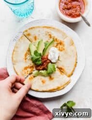 A hand grabbing a slice of a golden brown cheese quesadilla on a white plate that has been topped with avocado, sour cream, and salsa.