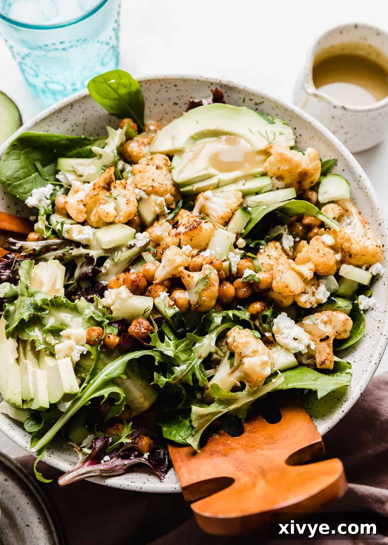 Wooden salad tongs in a large bowl that is full of mixed greens, roasted cauliflower, chickpeas, avocado, and cucumbers. 