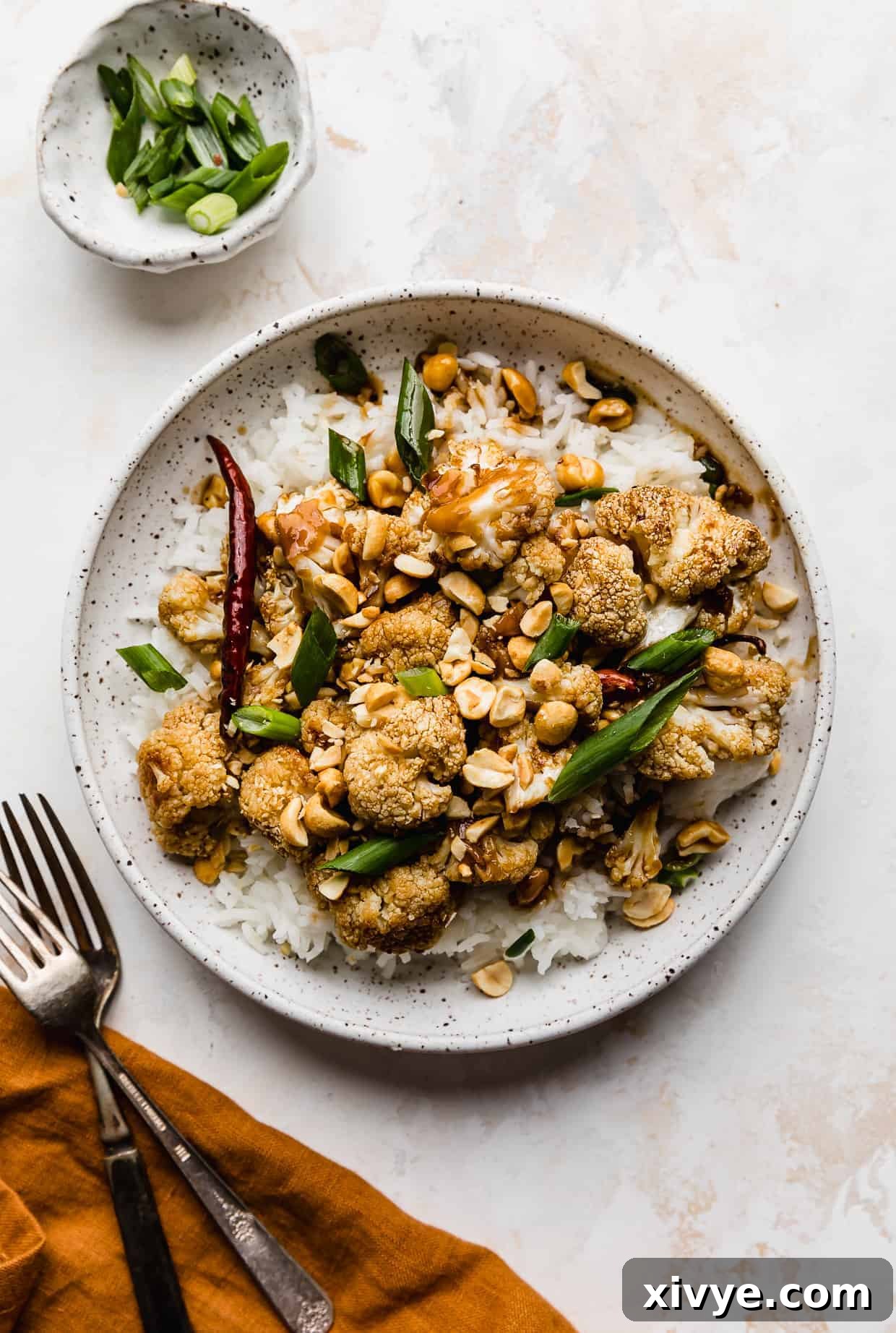 A white plate with green onion and peanut garnished Kung Pao Cauliflower, against a cream colored background.