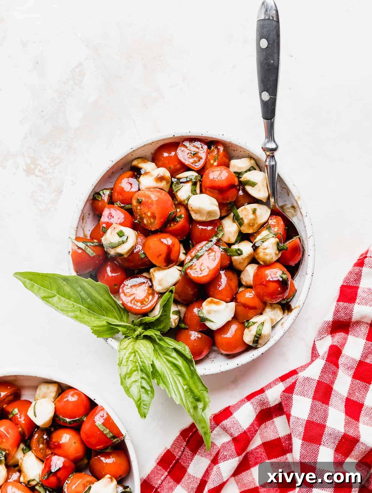 Vibrant Cherry Tomato Caprese Salad 8 A Cherry Tomato Caprese Salad in a white bowl with a sprig of fresh basil in it with a red checkered napkin next to the bowl.