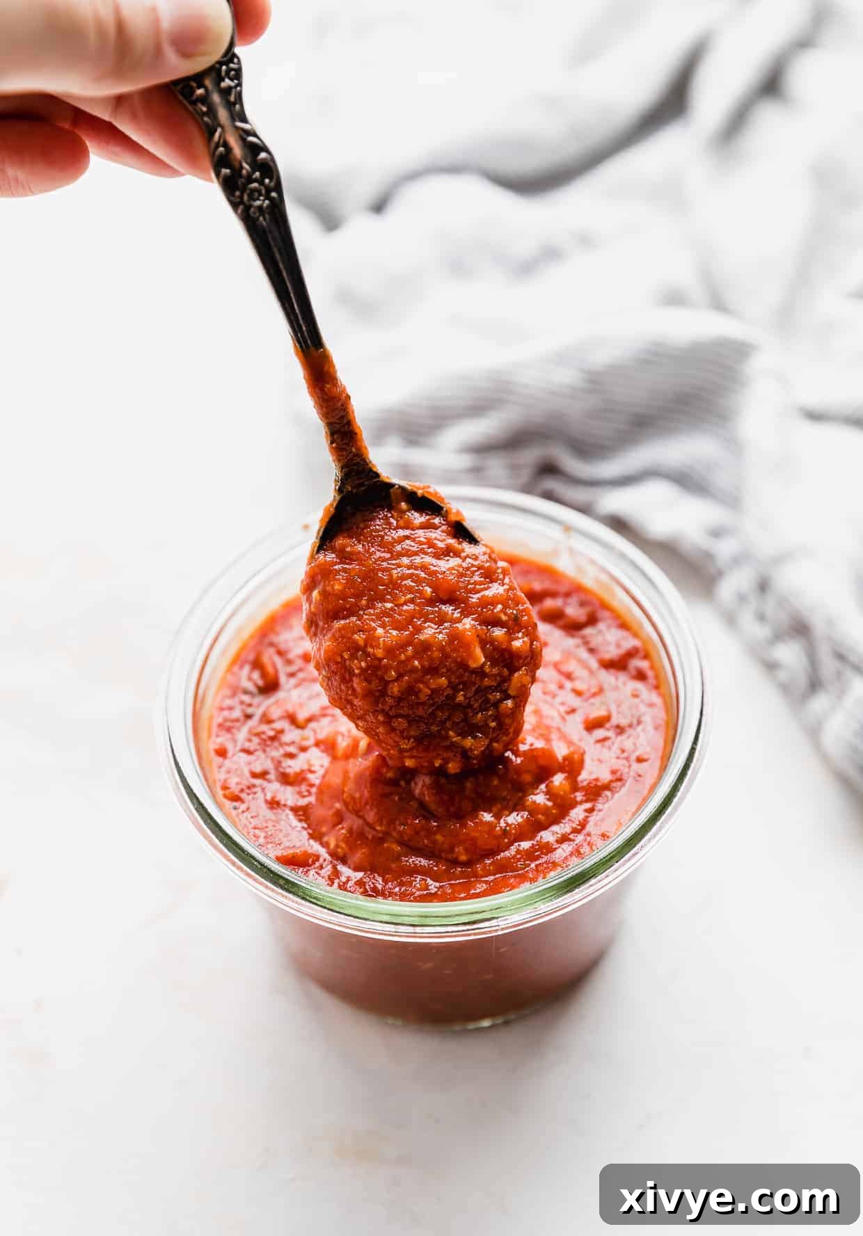 A spoon scooping up homemade pizza sauce from a glass jar, highlighting its rich, thick texture.