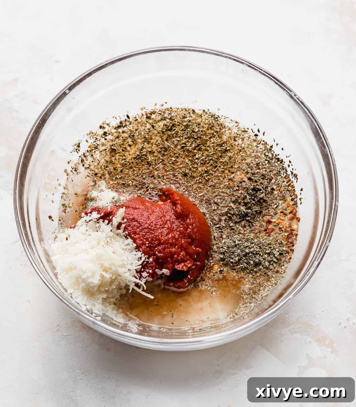 A glass bowl filled with all the ingredients for homemade tomato paste pizza sauce, ready to be mixed.