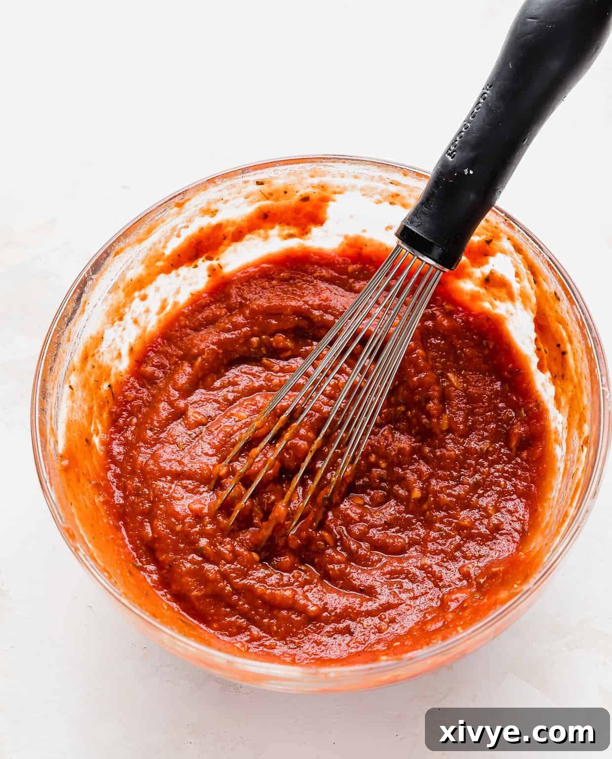 A whisk stirring homemade tomato paste pizza sauce in a glass bowl.
