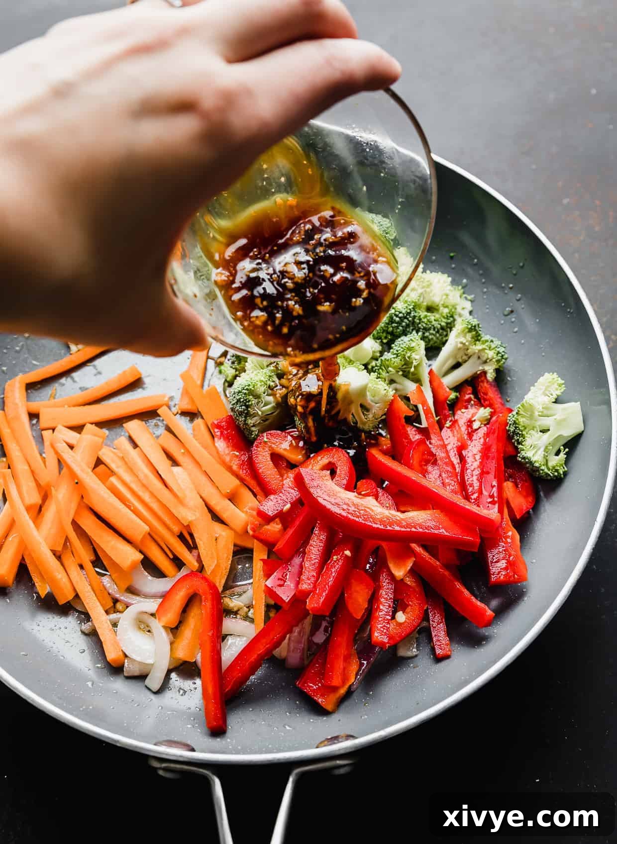 A hand pouring a brown sauce overtop of veggies in a skillet.