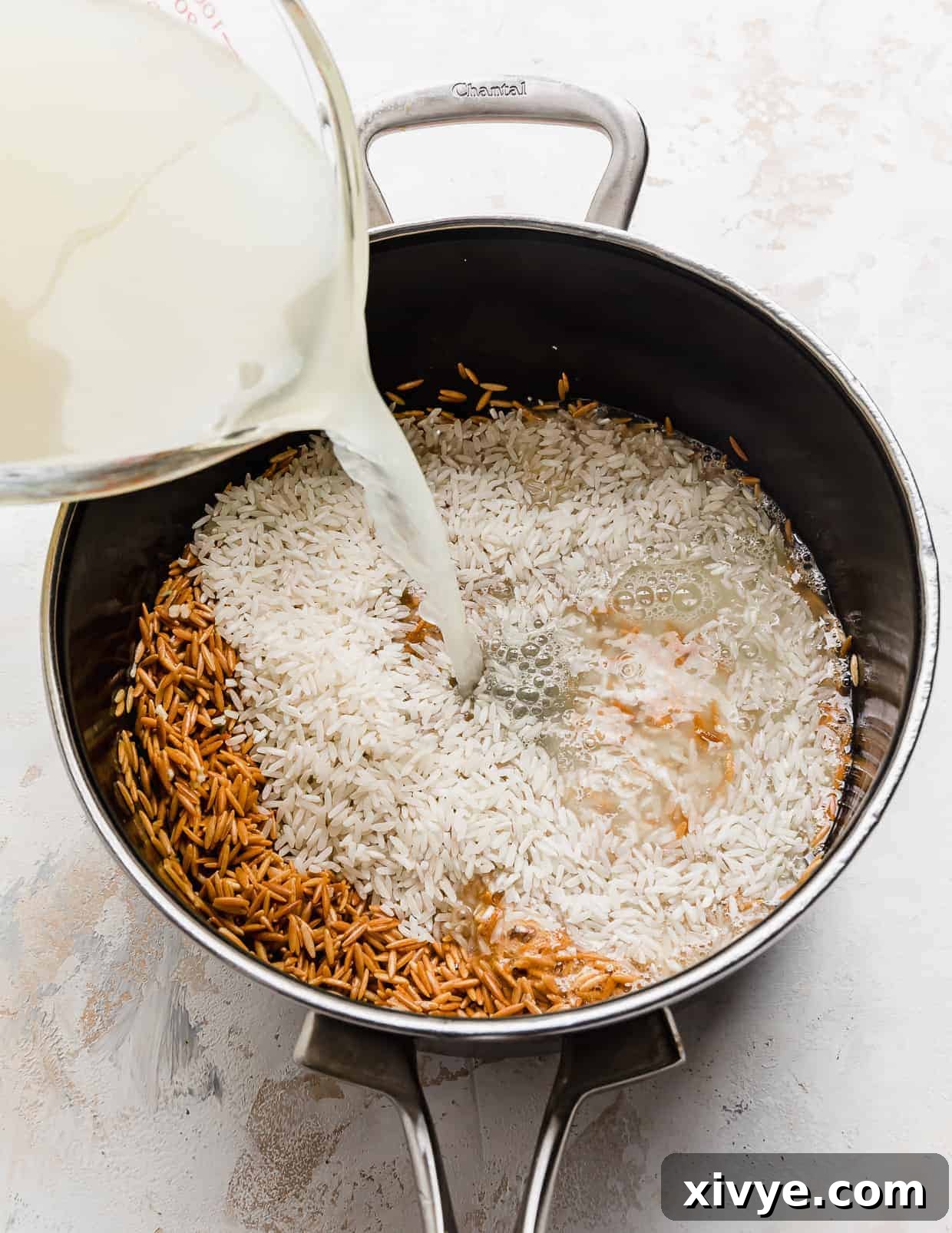 Orzo Pilaf 7 Rich chicken broth is being poured over the mixture of white rice and golden orzo in the saucepan, ready for simmering.