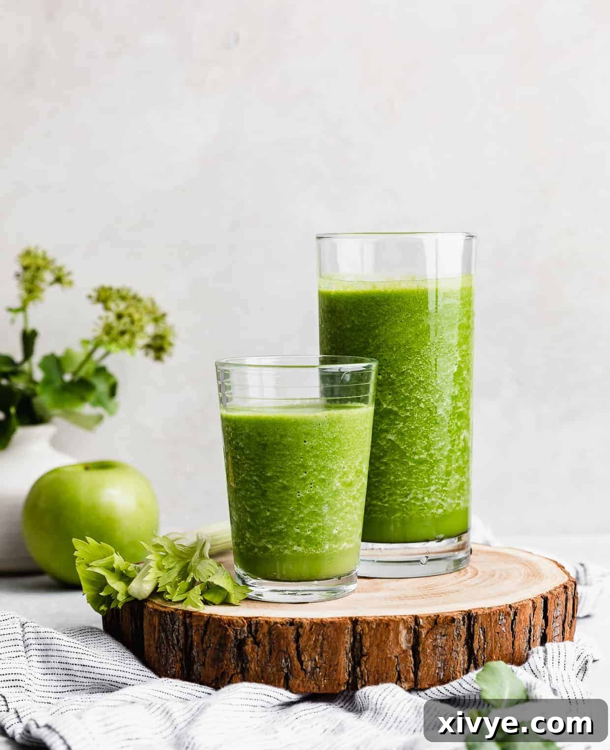Two glasses full of dark green celery smoothie on a wooden round board.