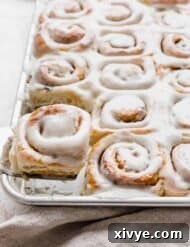 Irresistible Mini Cinnies 22 A metal spatula holding up a mini cinnamon roll in front of a baking sheet full of frosted cinnamon rolls.