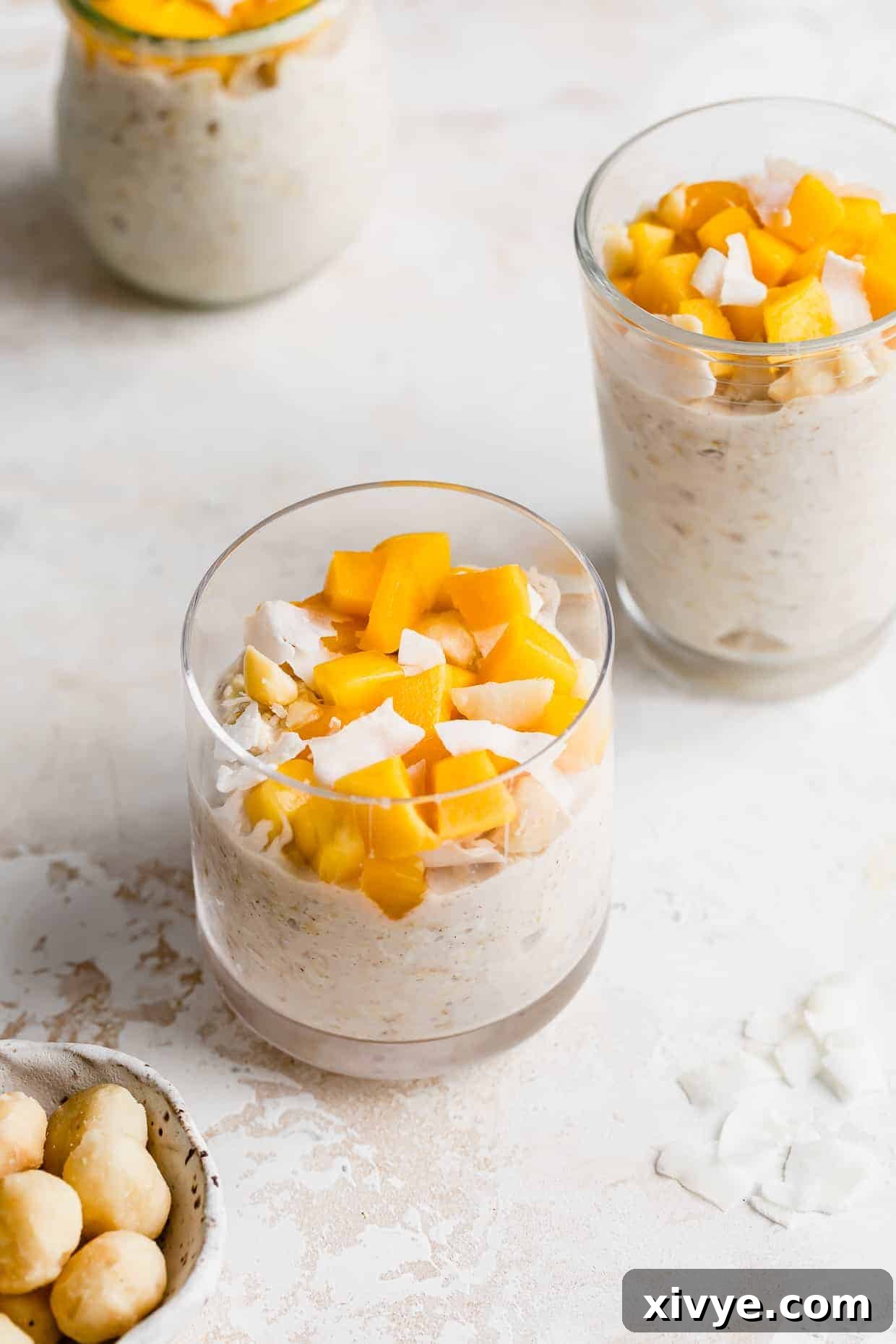 A clear cup full of Mango Overnight Oats topped with diced mangos and shaved coconut flakes, showcasing a vibrant and appealing breakfast.