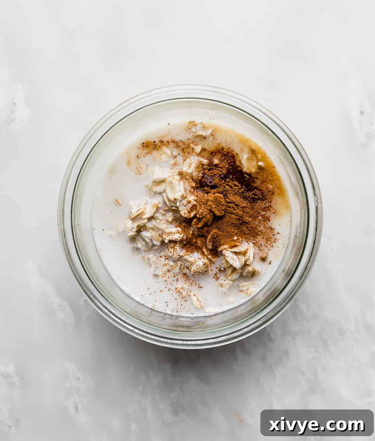 A glass jar filled with a mixture of rolled oats, milk, cinnamon, and nutmeg, ready to be chilled.