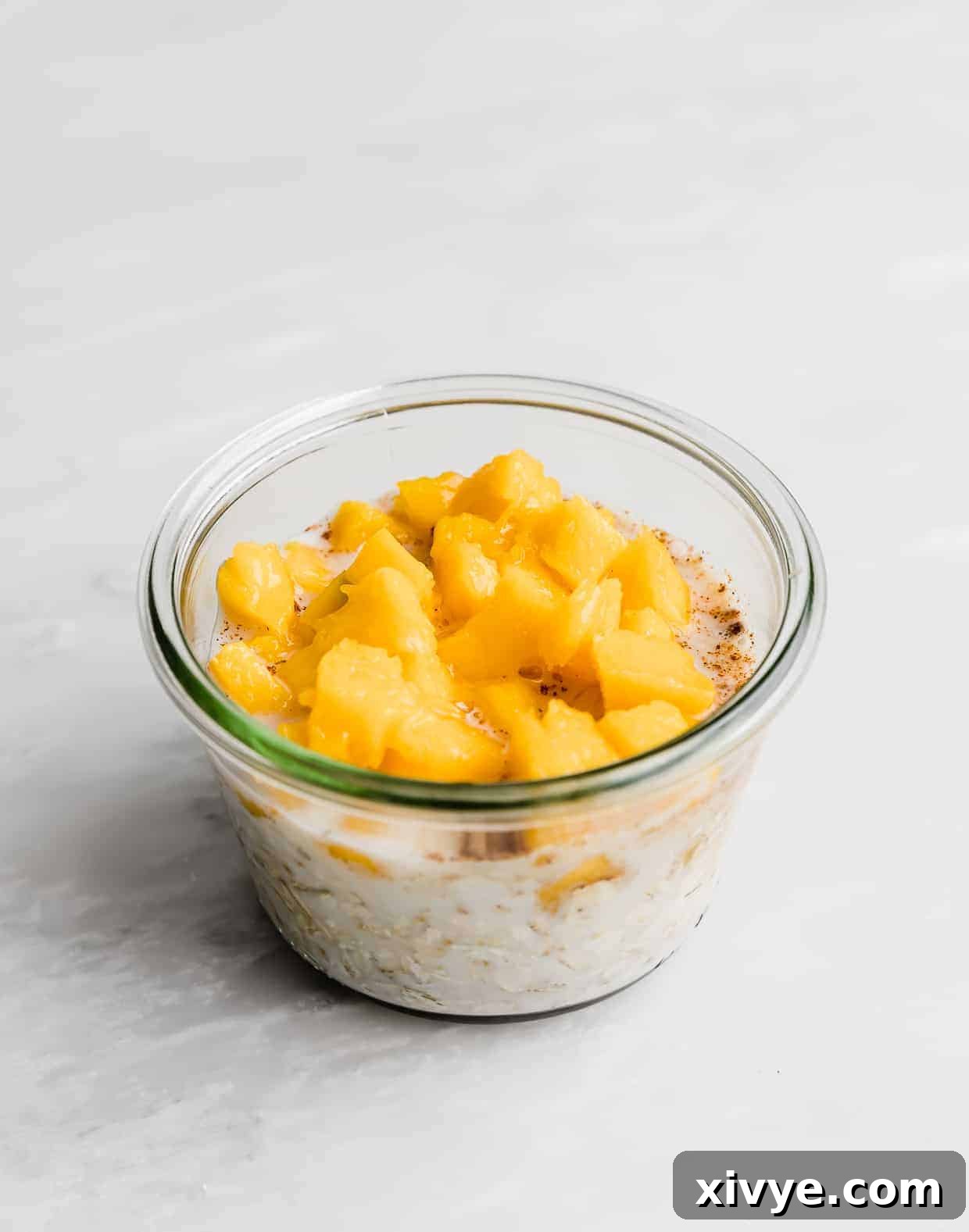 Diced mangos overtop of a milk and oat mixture in a glass jar, showing the final mix before chilling.