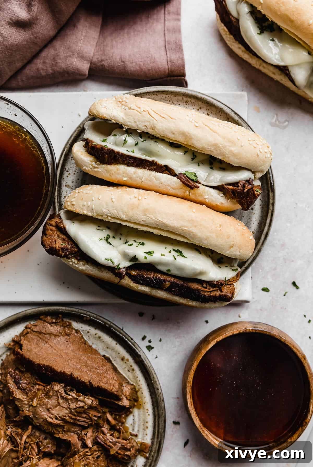 Ultimate Slow Cooker French Dip 2 Two Slow Cooker French Dip Sandwiches on a gray plate with two bowls of au jus beside it. The sandwiches are golden brown with melted provolone cheese and thinly sliced beef, inviting to dip.