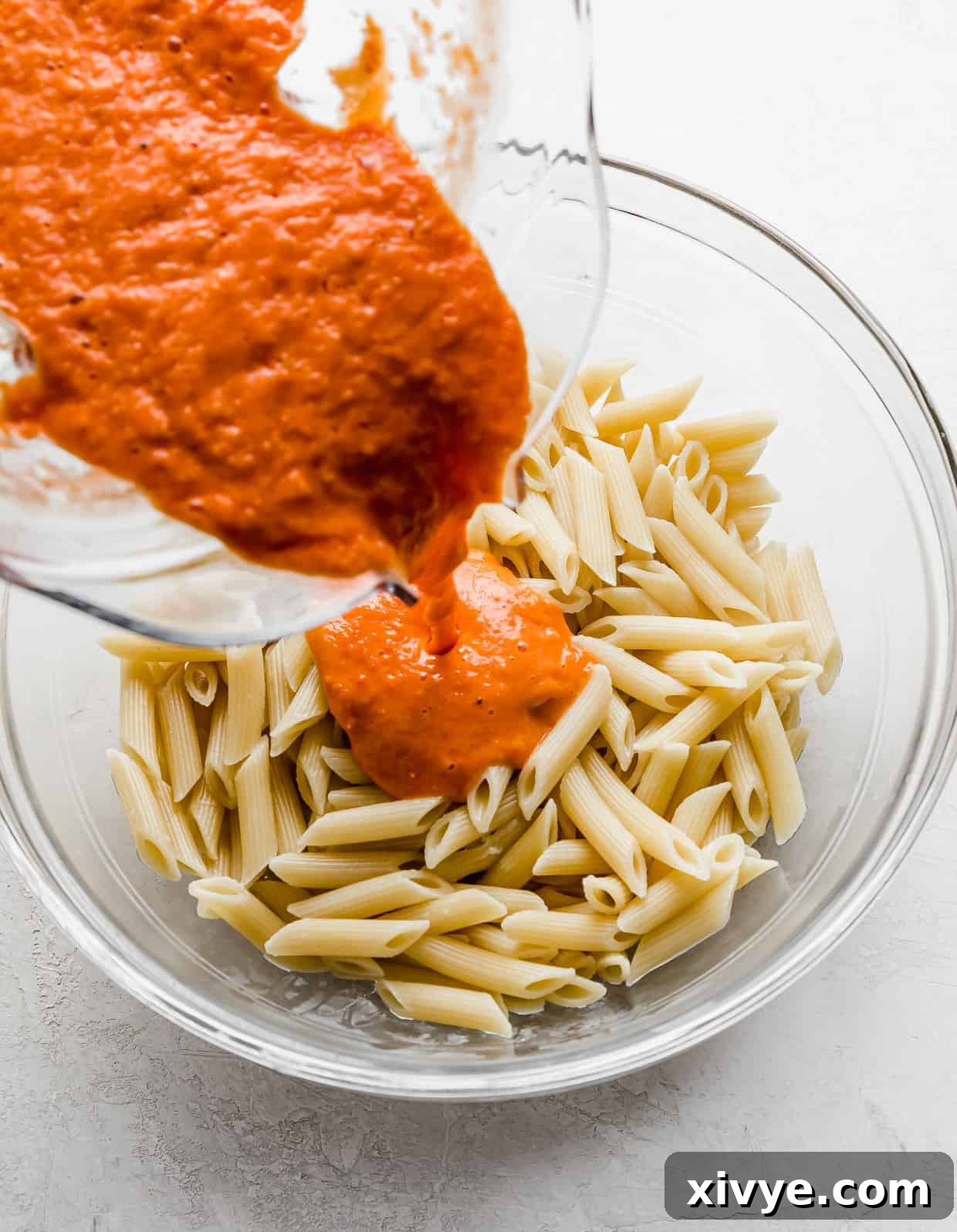 Fresh pureed tomato sauce being poured overtop cooked penne noodles in a glass bowl.