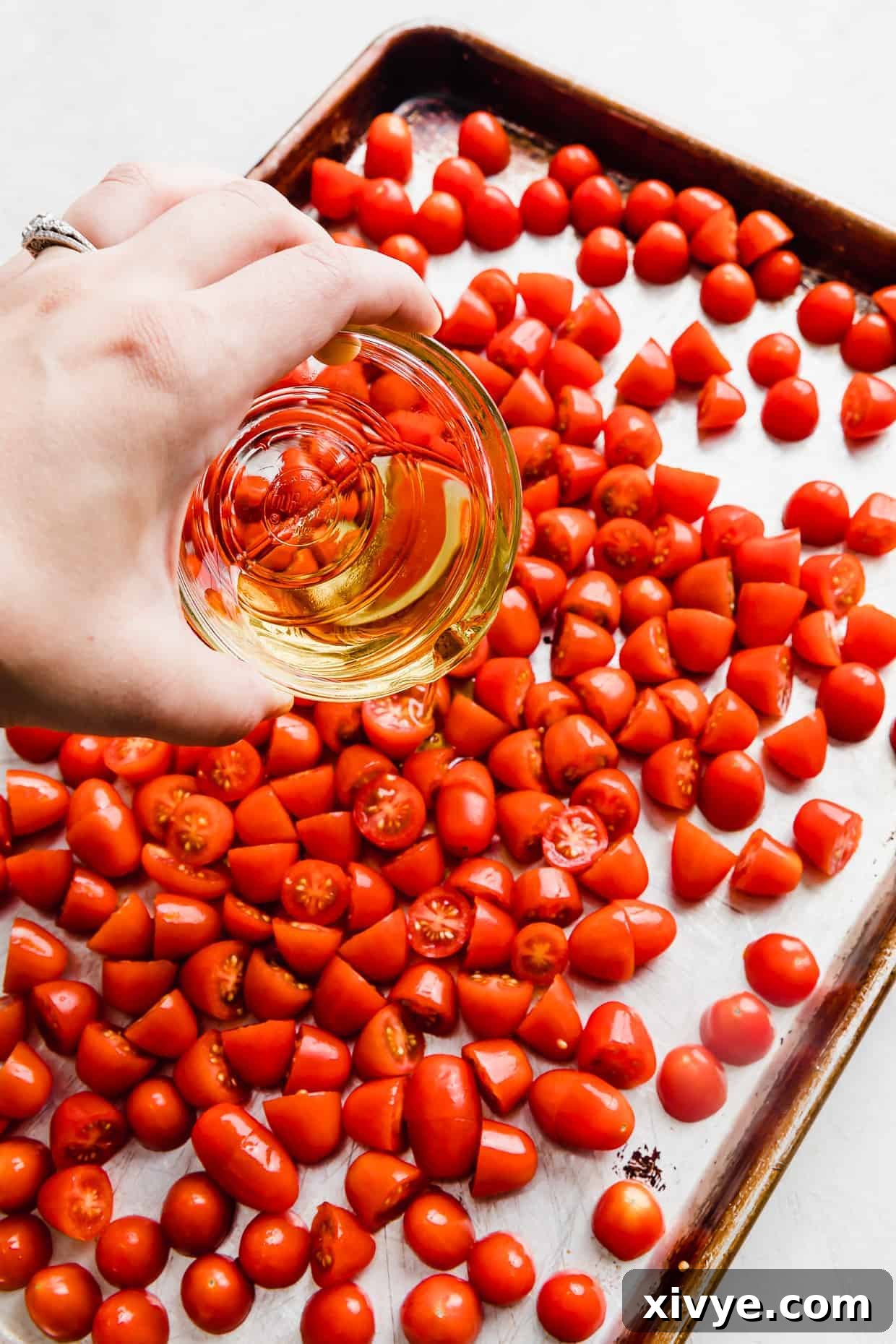 Olive oil in a glass bowl being drizzled over grape tomatoes on a baking sheet, in preparation to make roasted tomatoes. 